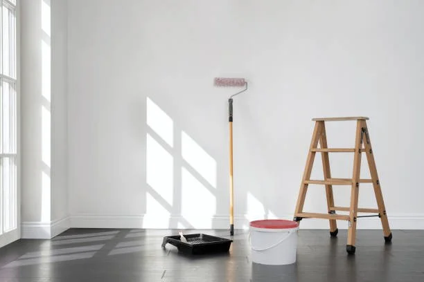 Empty room with a ladder, paint roller, paint tray, and paint bucket against a white wall.