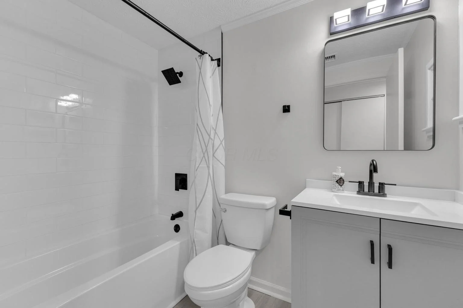 Modern bathroom with a white bathtub, black fixtures, toilet, and a vanity with a mirror and wall-mounted light