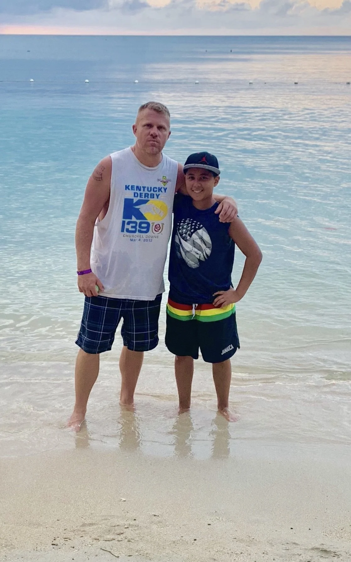 Two males standing in shallow water at a beach, smiling and posing for the photo. One is wearing a white sleeveless shirt with a Kentucky Derby logo, plaid shorts, and a purple wristband. The other is wearing a dark sleeveless shirt with an American flag design, rainbow-colored shorts, and a cap. The ocean and sky are visible in the background.