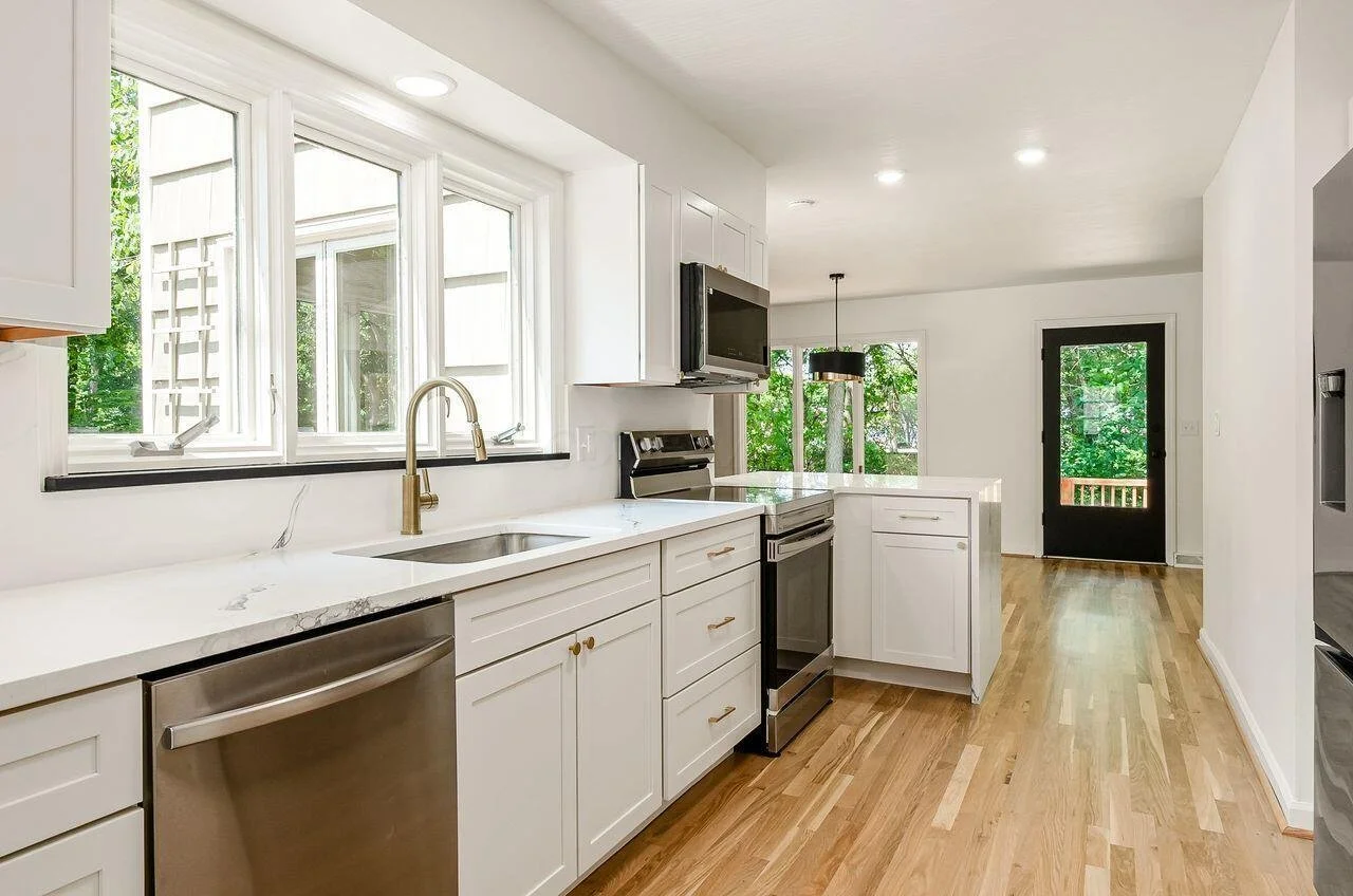A bright, modern kitchen with white cabinets, a marble countertop, stainless steel appliances, and large windows overlooking greenery.