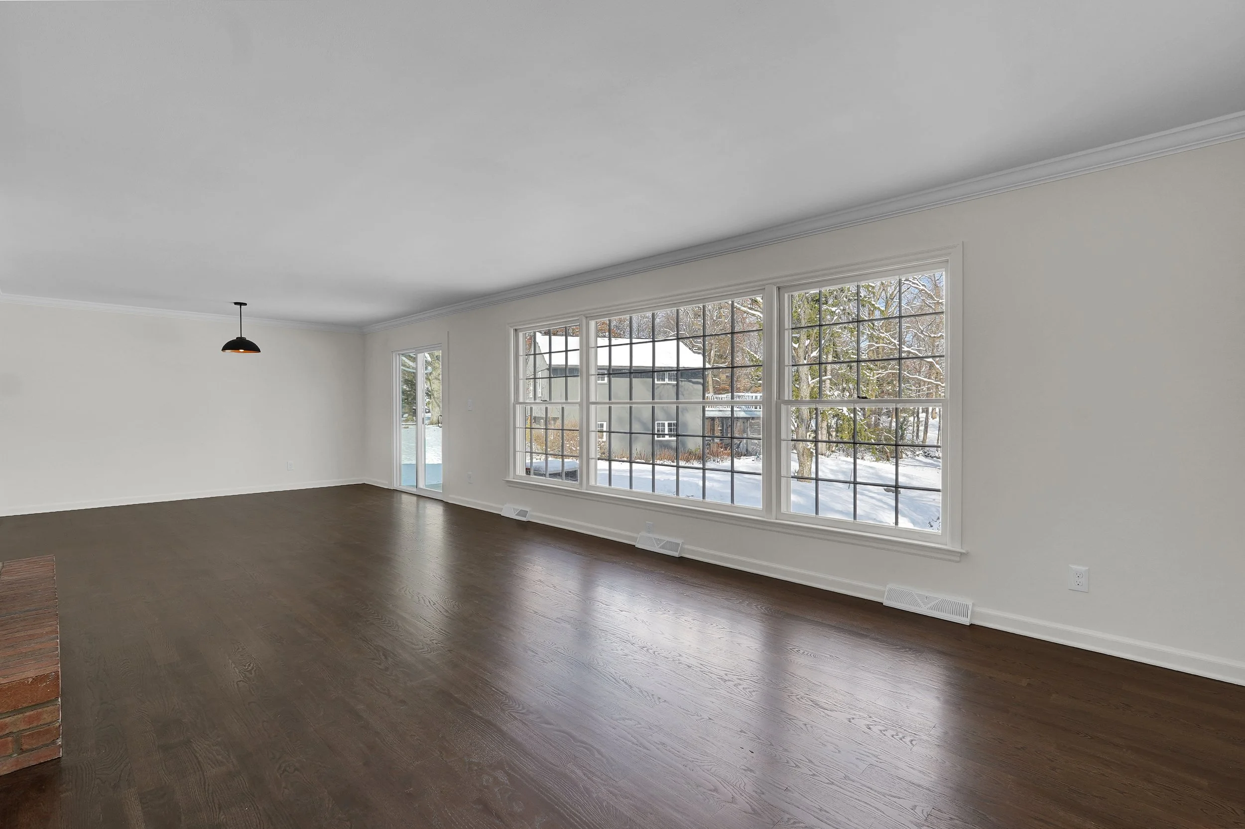 Empty living room with large windows showing a snowy outdoor scene and dark hardwood floors.