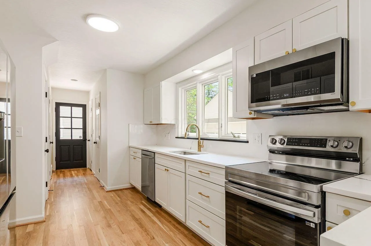 Kitchen with white cabinets, a window above the sink, stainless steel oven and microwave, and light wooden flooring.