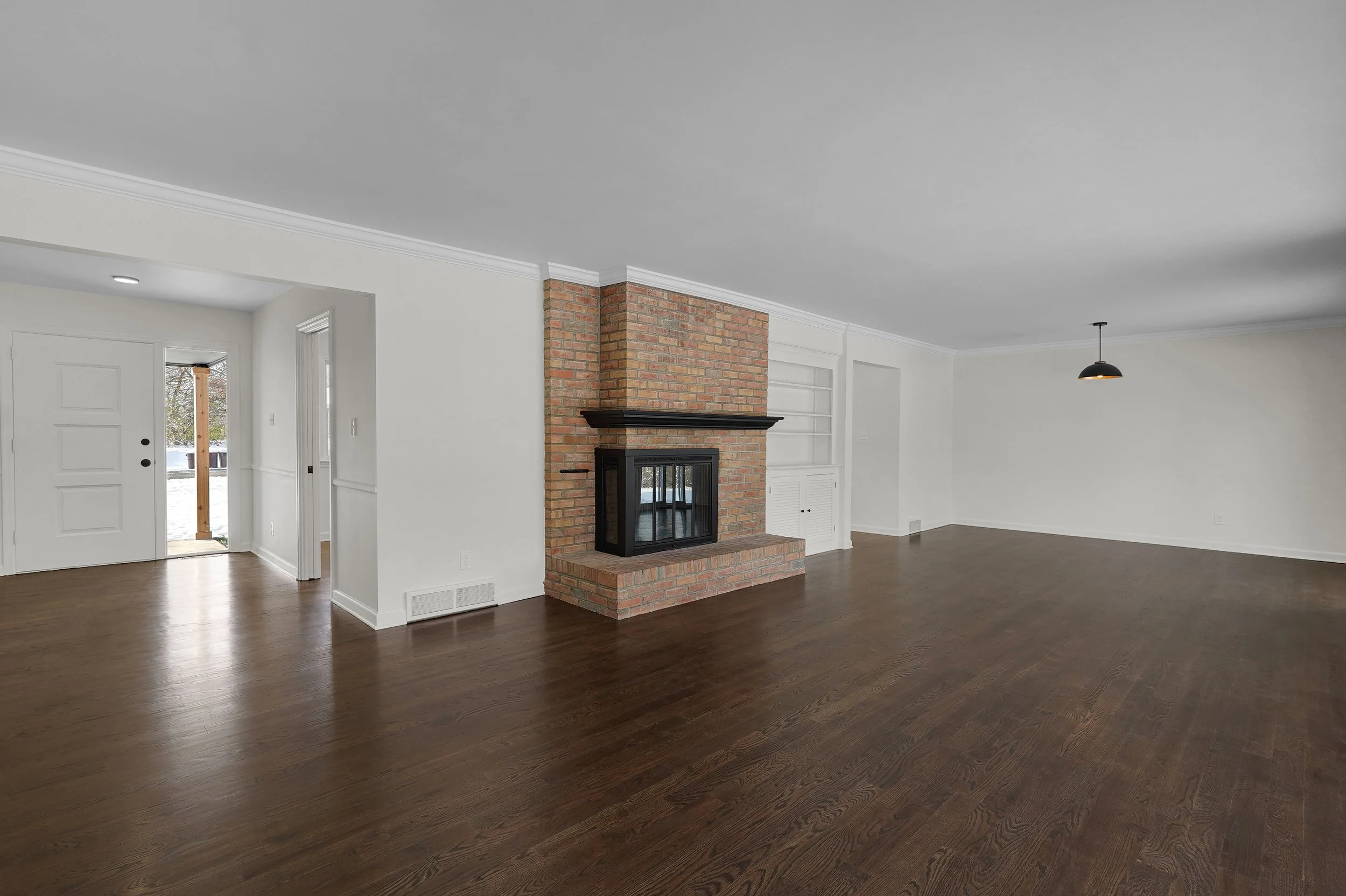 Empty living room with white walls, hardwood floors, brick fireplace, and large windows letting in natural light.