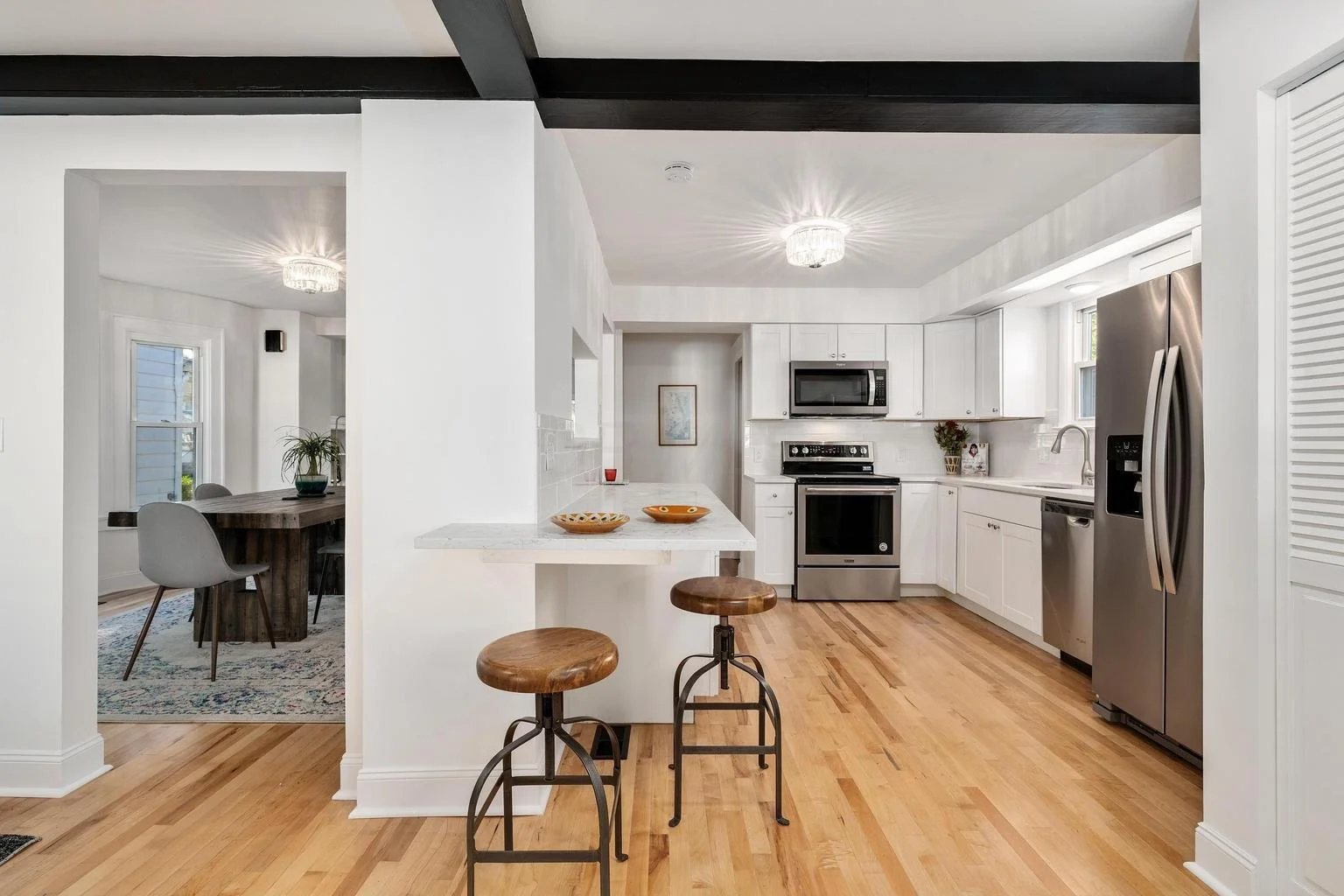 Modern kitchen with white cabinets, stainless steel appliances, hardwood floors, and a small breakfast bar with two wooden stools.