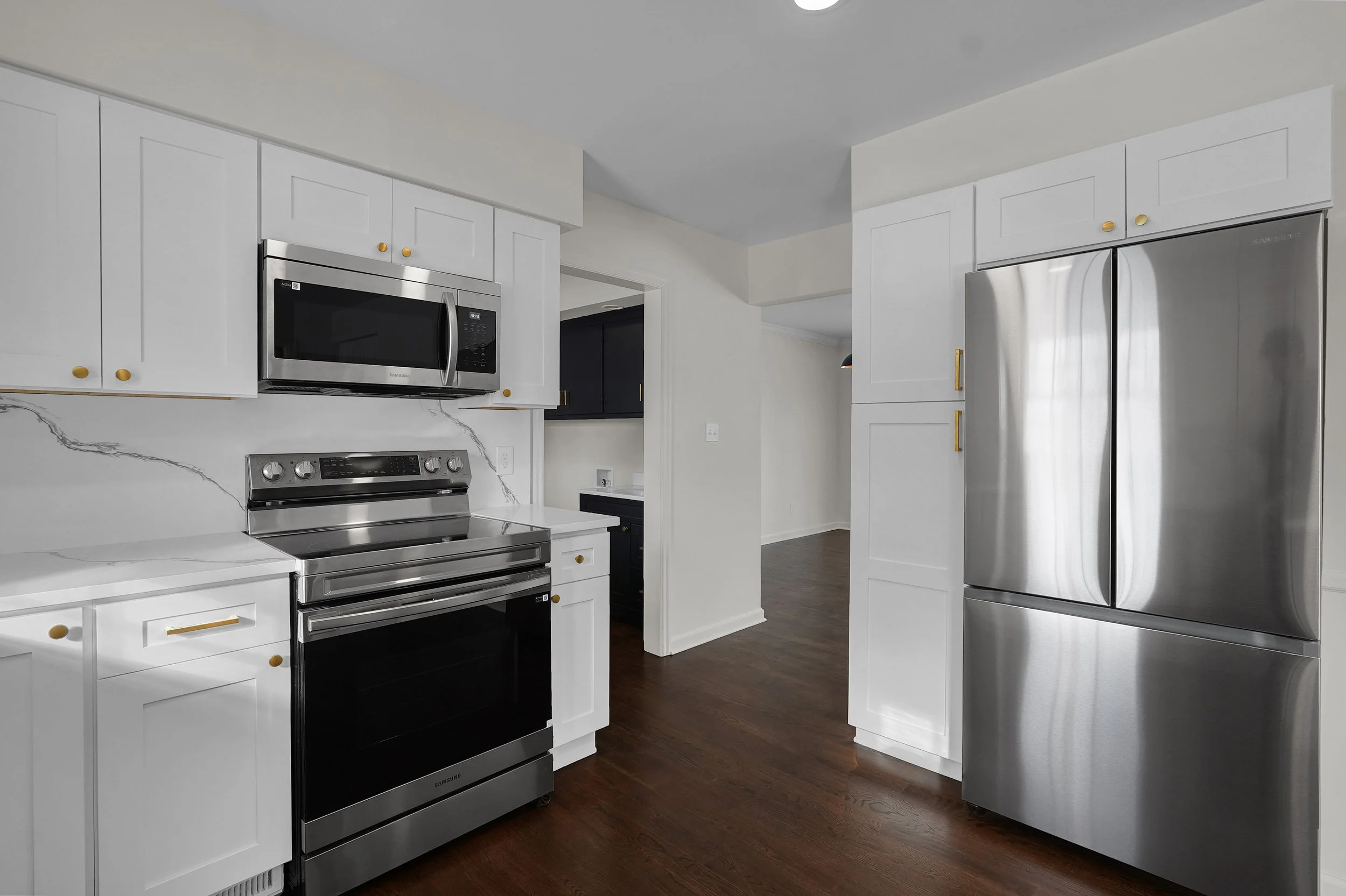 Modern kitchen with white cabinetry, stainless steel microwave, oven, and refrigerator, dark hardwood floors.