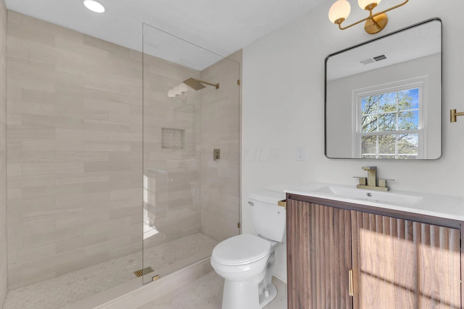 Modern bathroom with glass-enclosed shower, white toilet, wooden vanity with a white sink, large mirror, and a window showing blue sky and tree outside.
