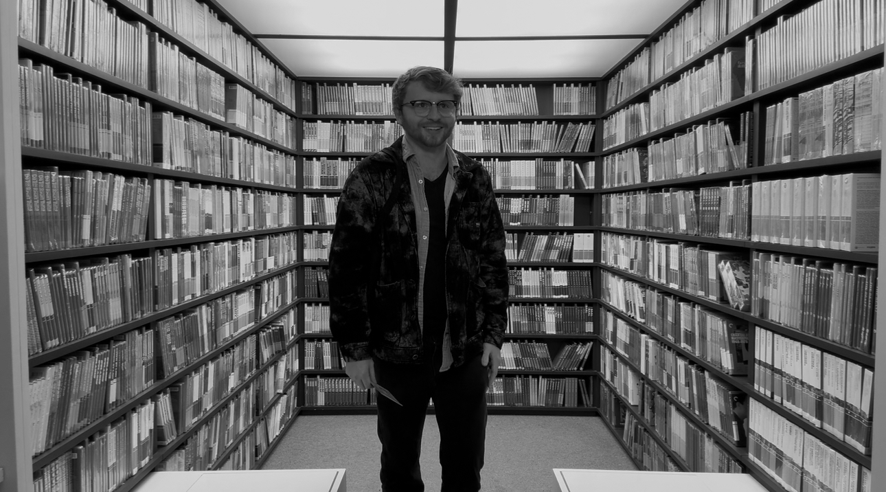 A man standing in a library aisle surrounded by tall bookshelves filled with books.