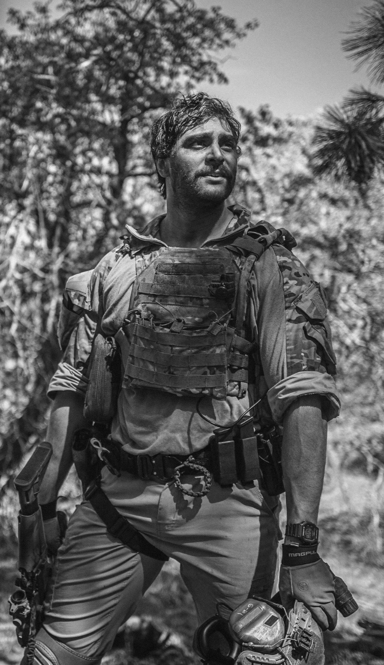 A man dressed in military or tactical gear outdoors, holding a gun, surrounded by trees.
