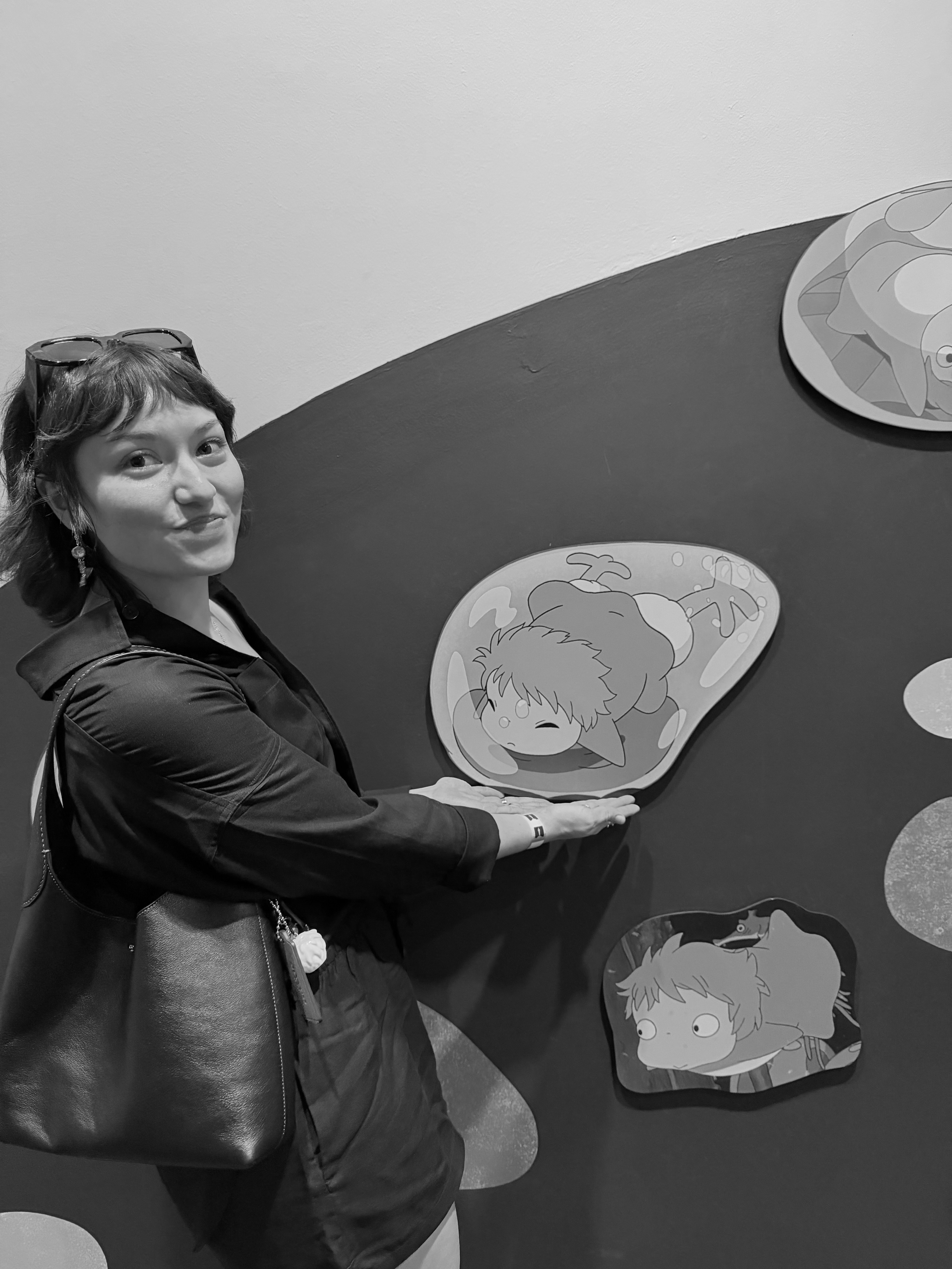 A woman with glasses on her head, earrings, and a dark blazer, holding her hand under a cartoon character illustration on a wall. The cartoon shows a girl with short hair, a hat, and a cape, lying down with a pained expression. The wall has additional cartoon illustrations, including a girl with wide eyes and an animal with a long snout.