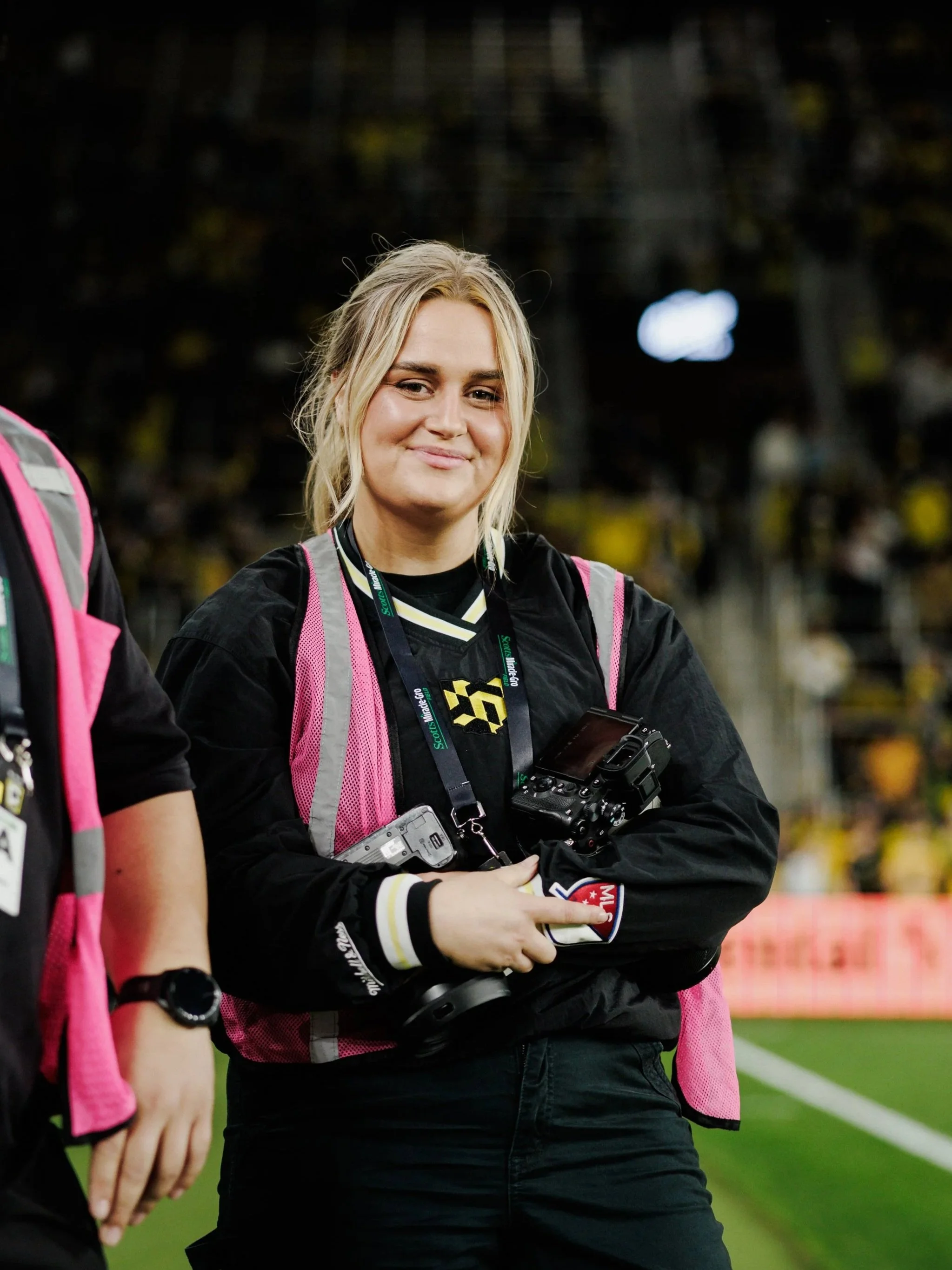 Young woman standing on a sports field, holding a camera and wearing a photographer's lanyard and camera gear around her neck, smiling at the camera.