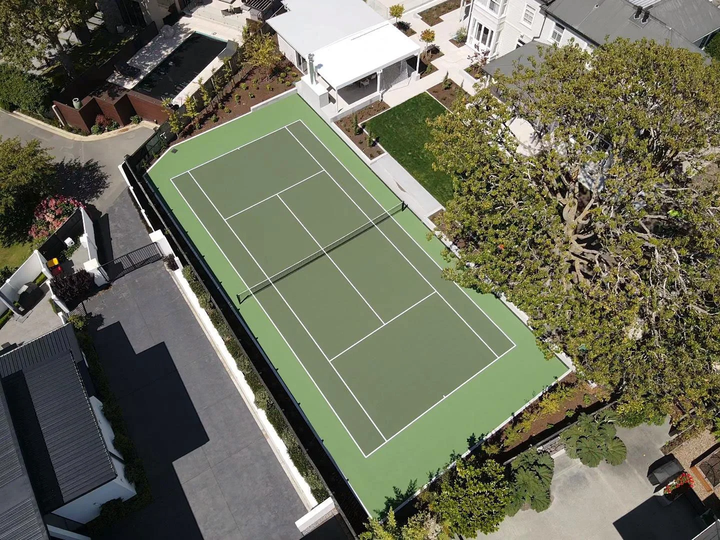 Aerial view of a green tennis court with white lines, surrounded by trees, houses, and a driveway in a residential neighborhood.