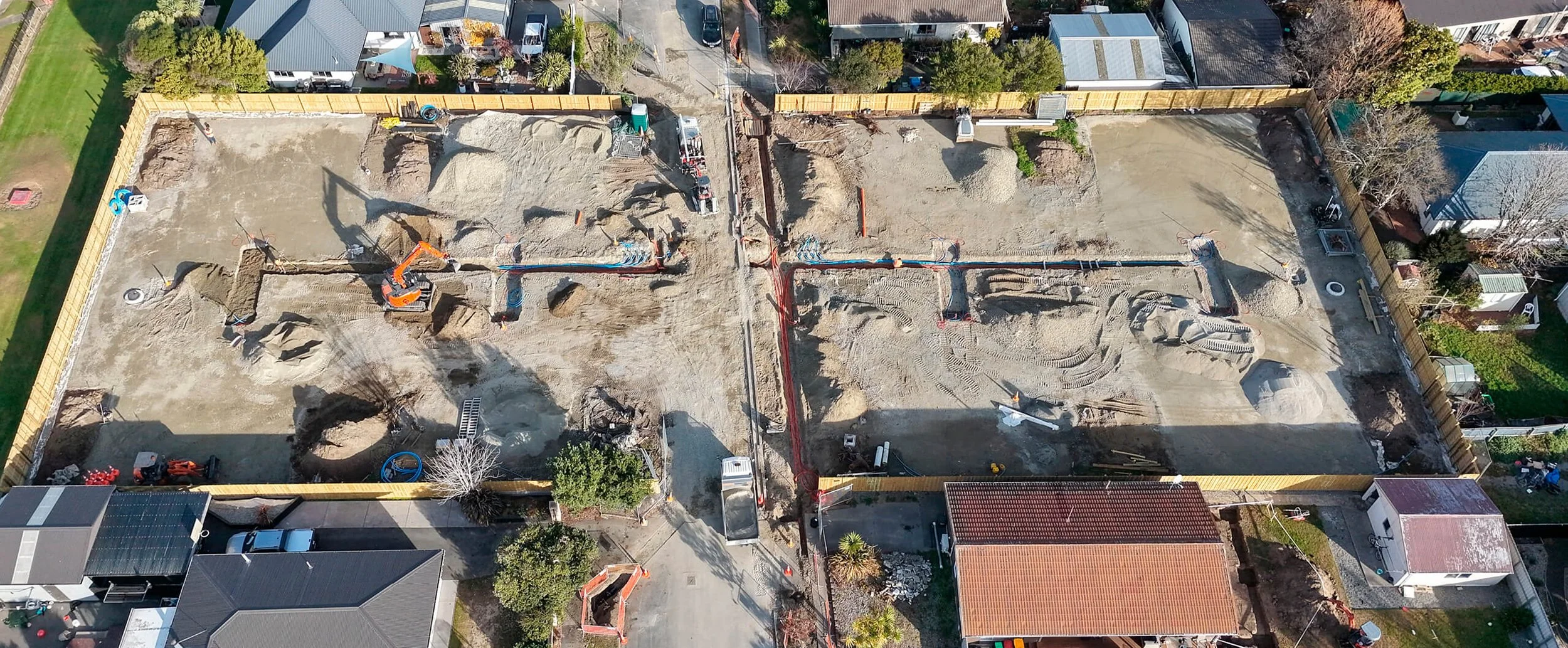 Aerial view of a construction site with excavators, trenches, and dirt piles, surrounded by residential houses and fences.