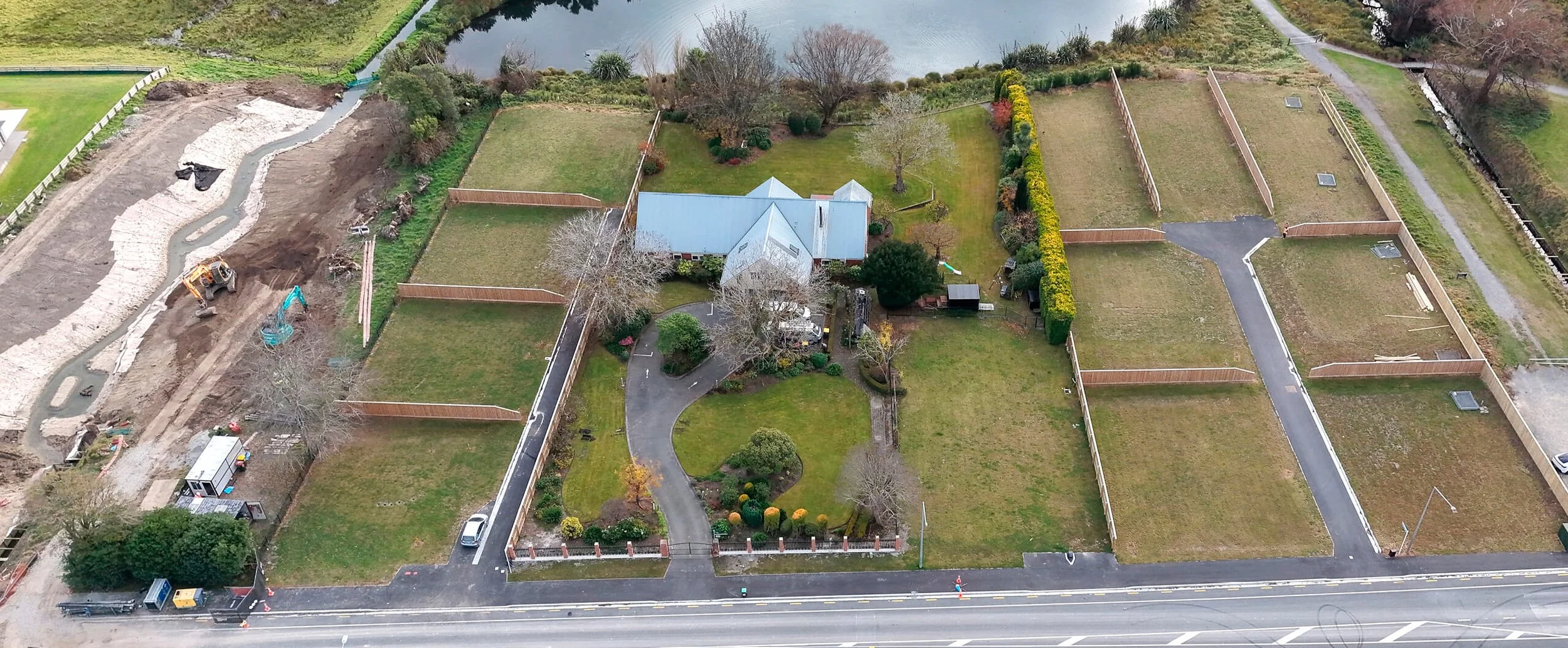 An aerial view of a property with a house, gardens, fenced yards, and a small water body, with what appears to be construction work on the left side.