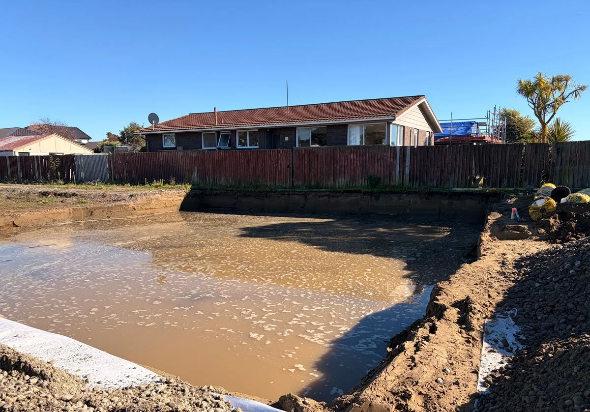 Construction site with a large cleared area filled with muddy water and dirt piles, with a house and trees in the background under a clear blue sky.