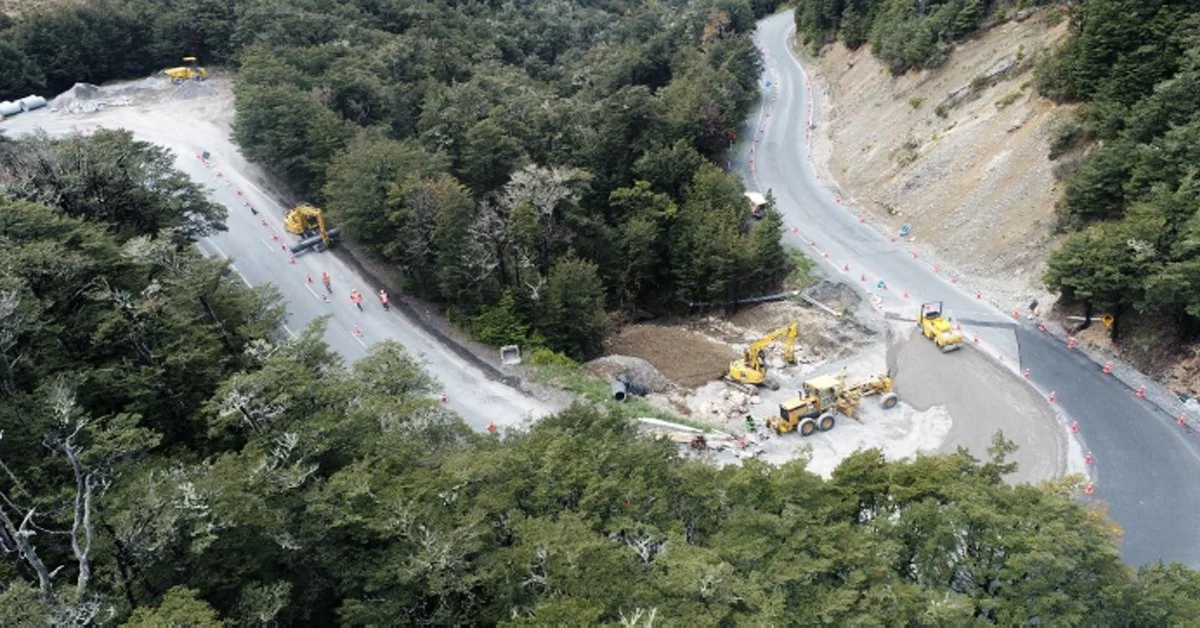 Construction workers and machinery repairing a winding mountain road surrounded by trees.