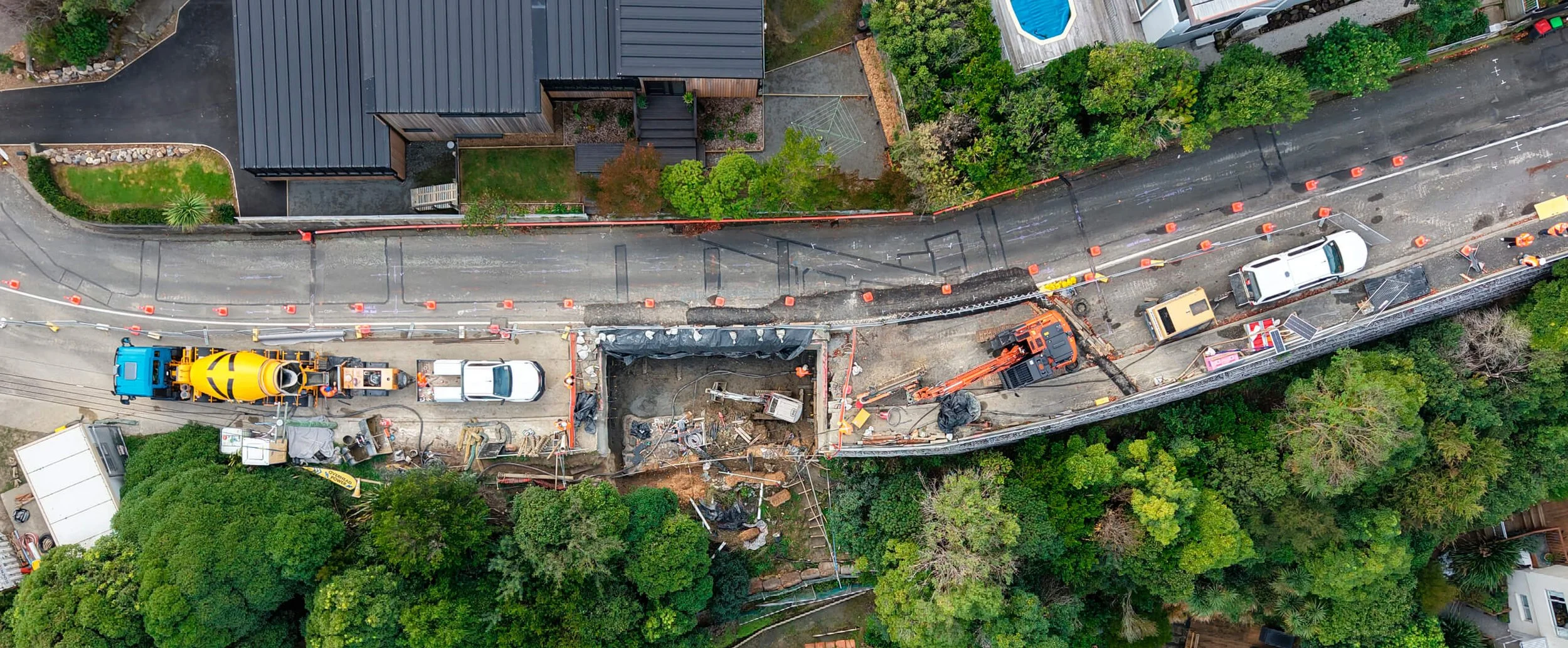Aerial view of road construction with orange cones, construction vehicles, and workers. Surrounding greenery and residential buildings.