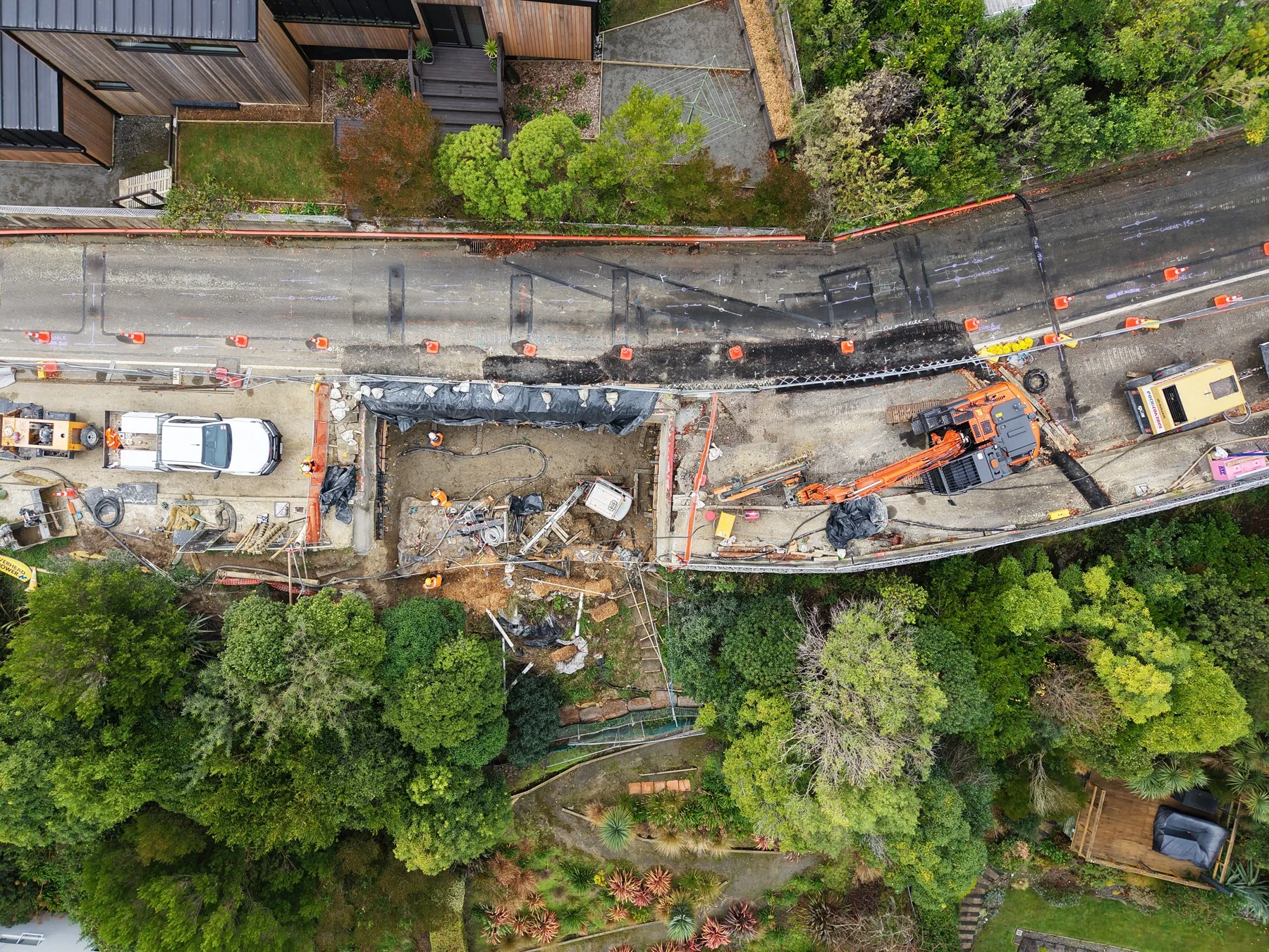 Aerial view of a construction site with machinery, workers, and orange safety cones on a partially excavated area surrounded by trees and residential buildings.