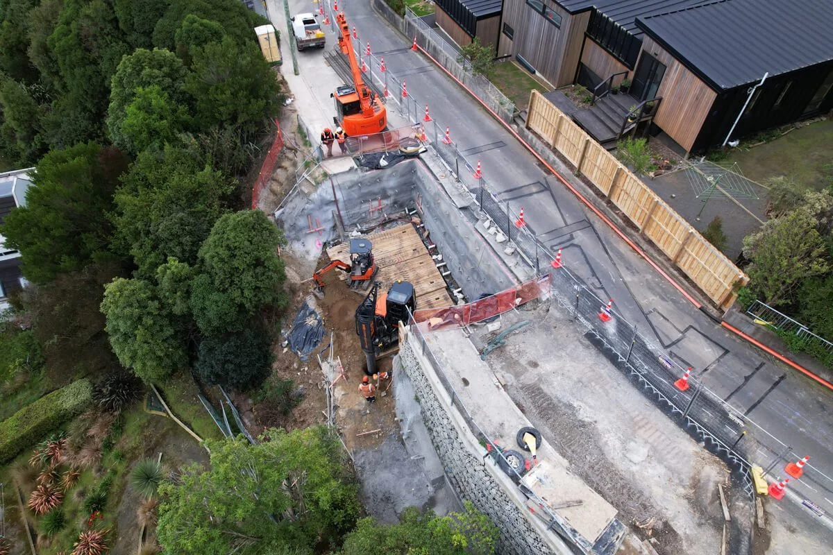 Construction site on a residential street with excavators, workers, orange safety cones, and temporary fencing, paving or repairing the road.