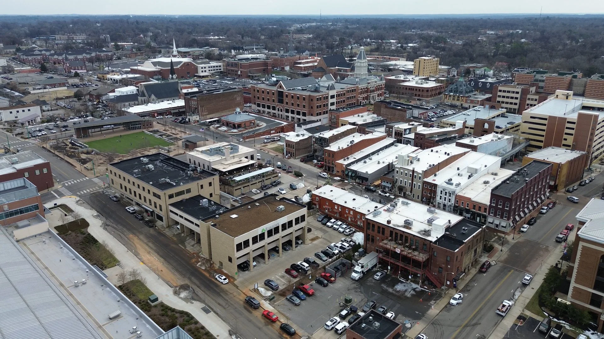 Aerial view of a downtown area with various commercial and residential buildings, parking lots, and a small park with a playground and green field.