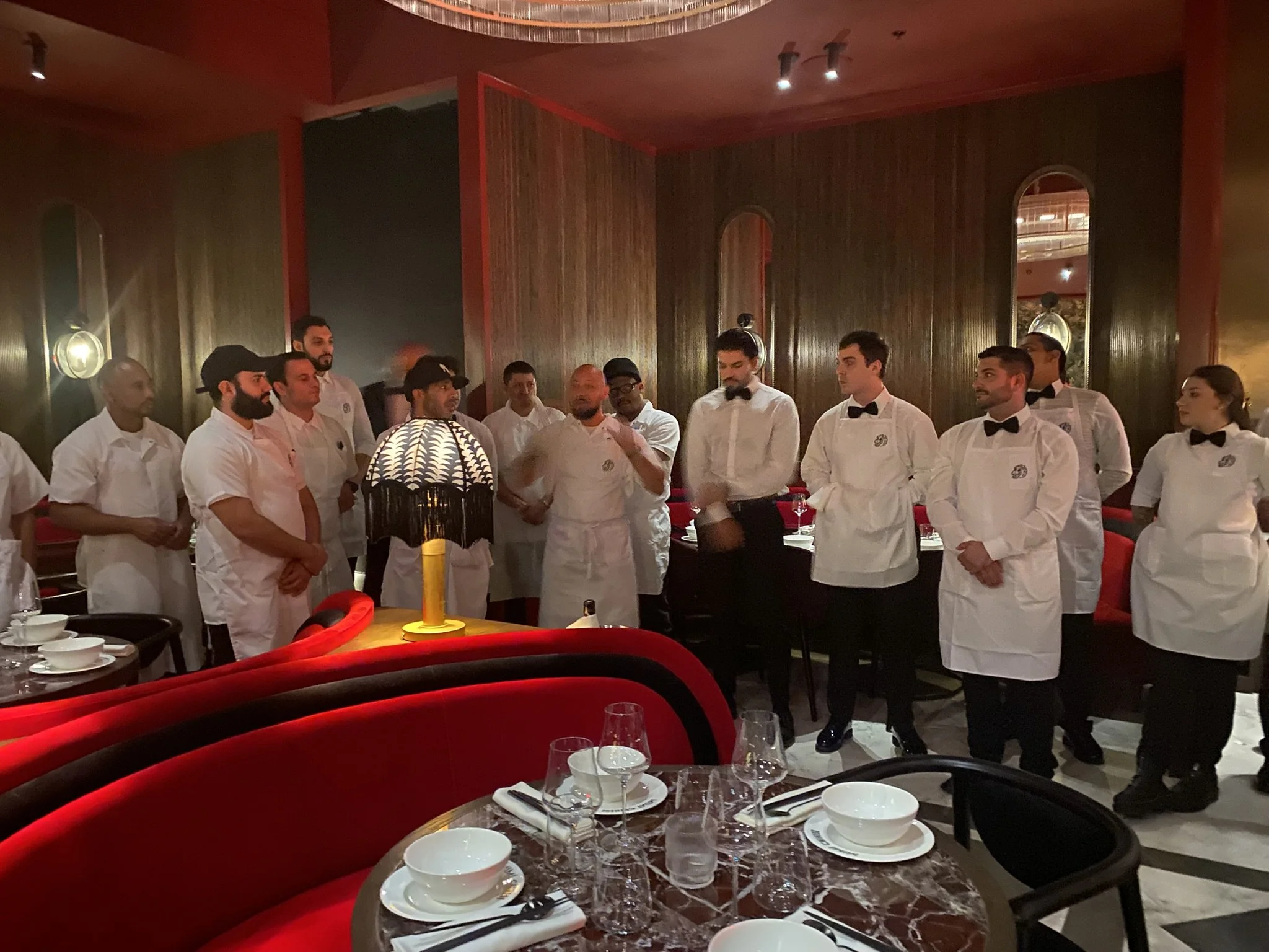 A group of restaurant staff, including chefs and waiters, standing together in a dimly lit dining area, preparing for a presentation or briefing.