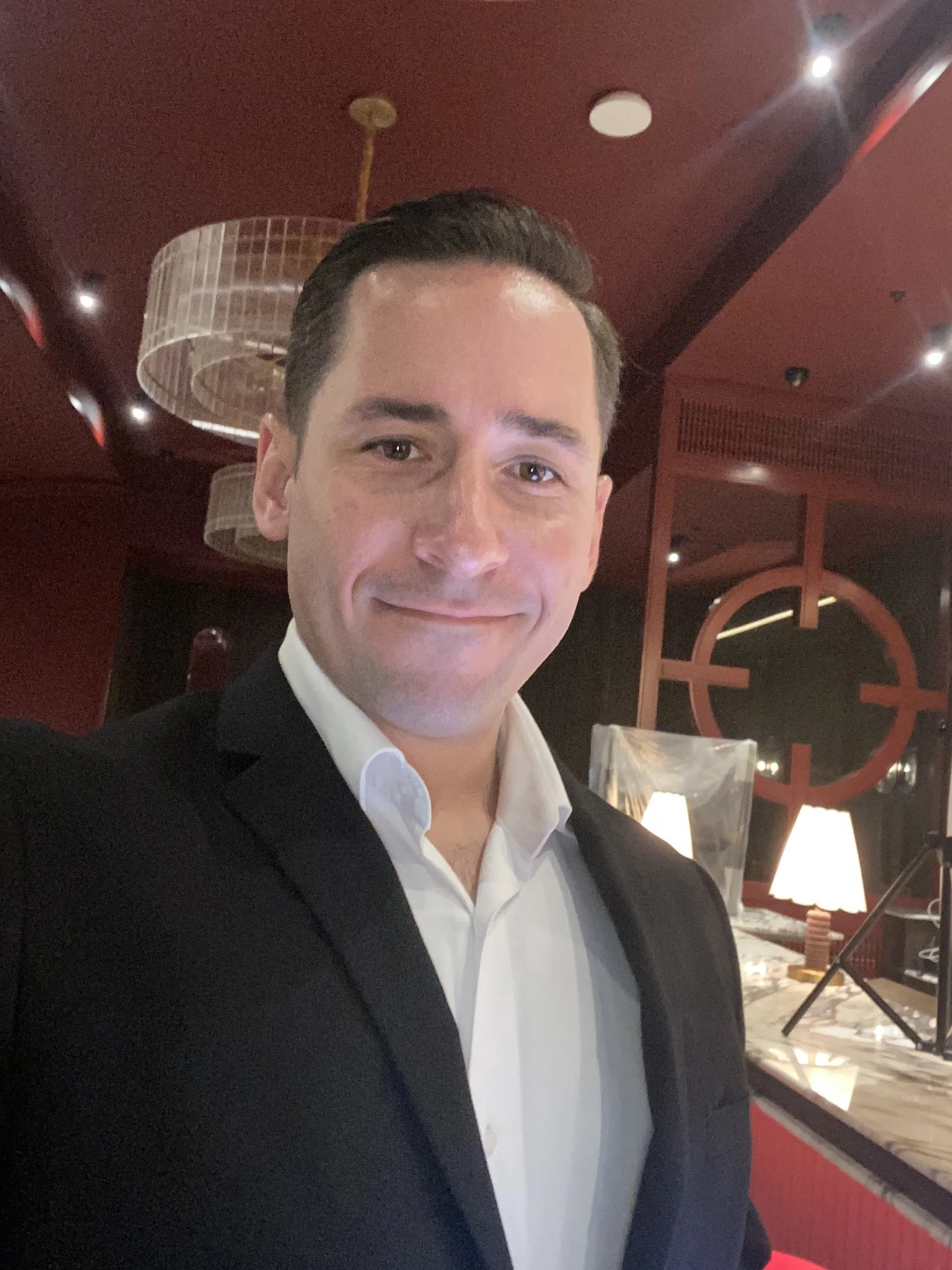 A man dressed in a formal black jacket and white shirt taking a selfie in an elegant room with dim lighting, lamps, and decorative elements.