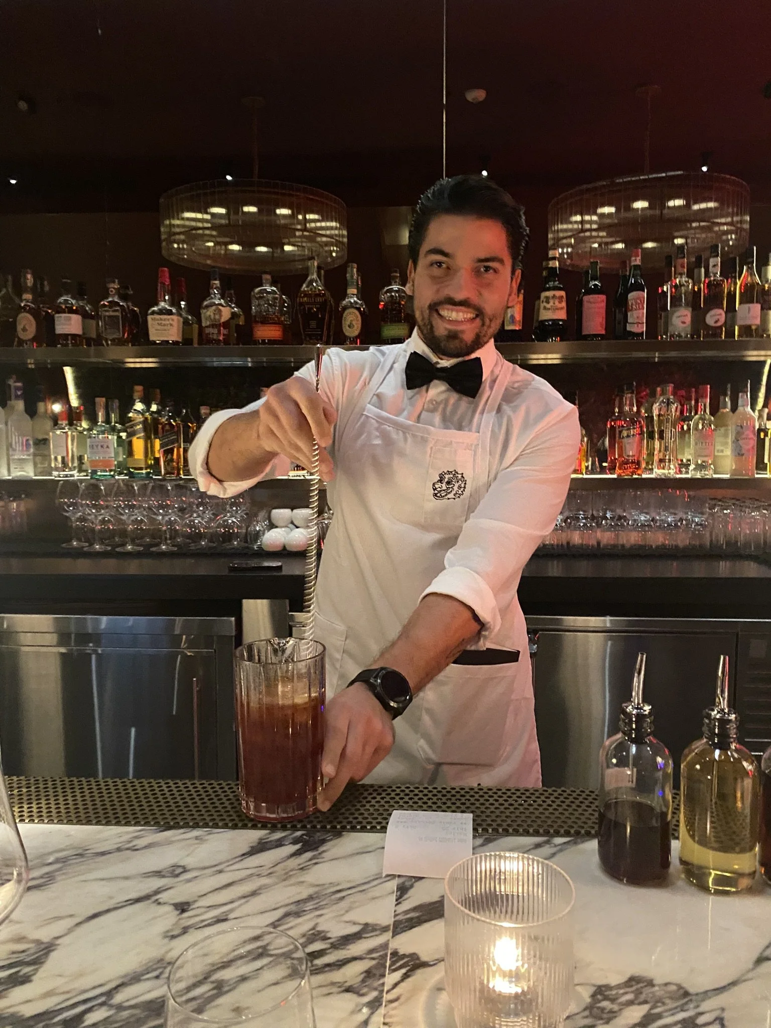 Bartender smiling in a tuxedo with a bowtie making a cocktail at a marble bar counter, with liquor bottles behind him.