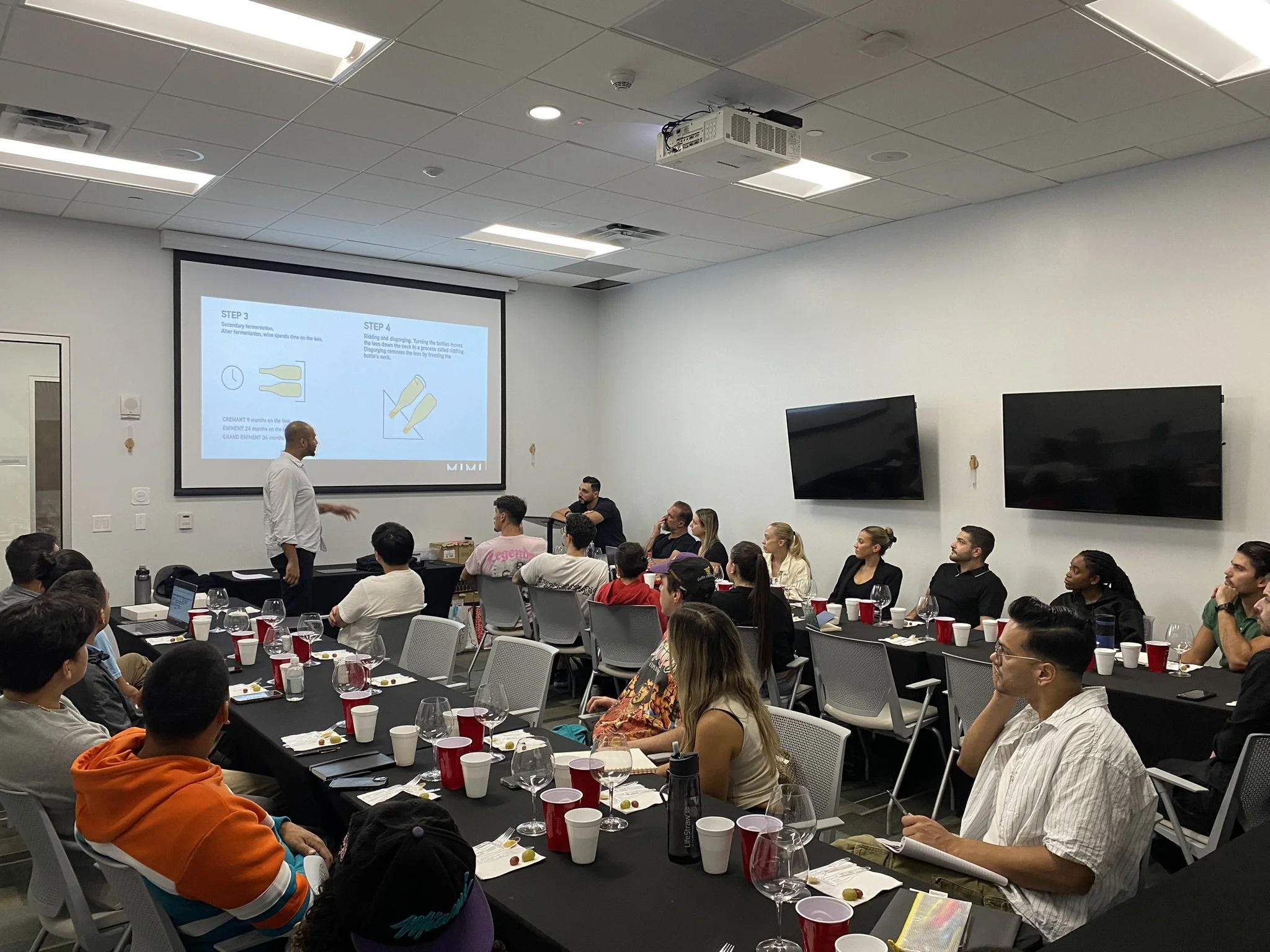 A group of people attending a presentation in a conference room with a speaker at the front, a large screen displaying slides, and multiple television monitors on the wall.