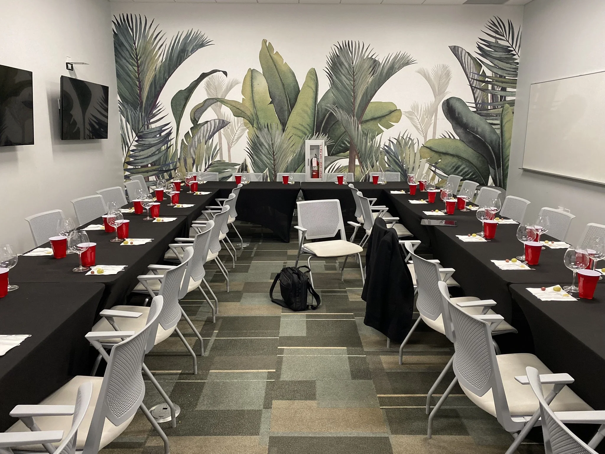Conference room with U-shaped black tables set with white napkins, wine glasses, and red cups, surrounded by white chairs, with a tropical leaf mural on the wall.