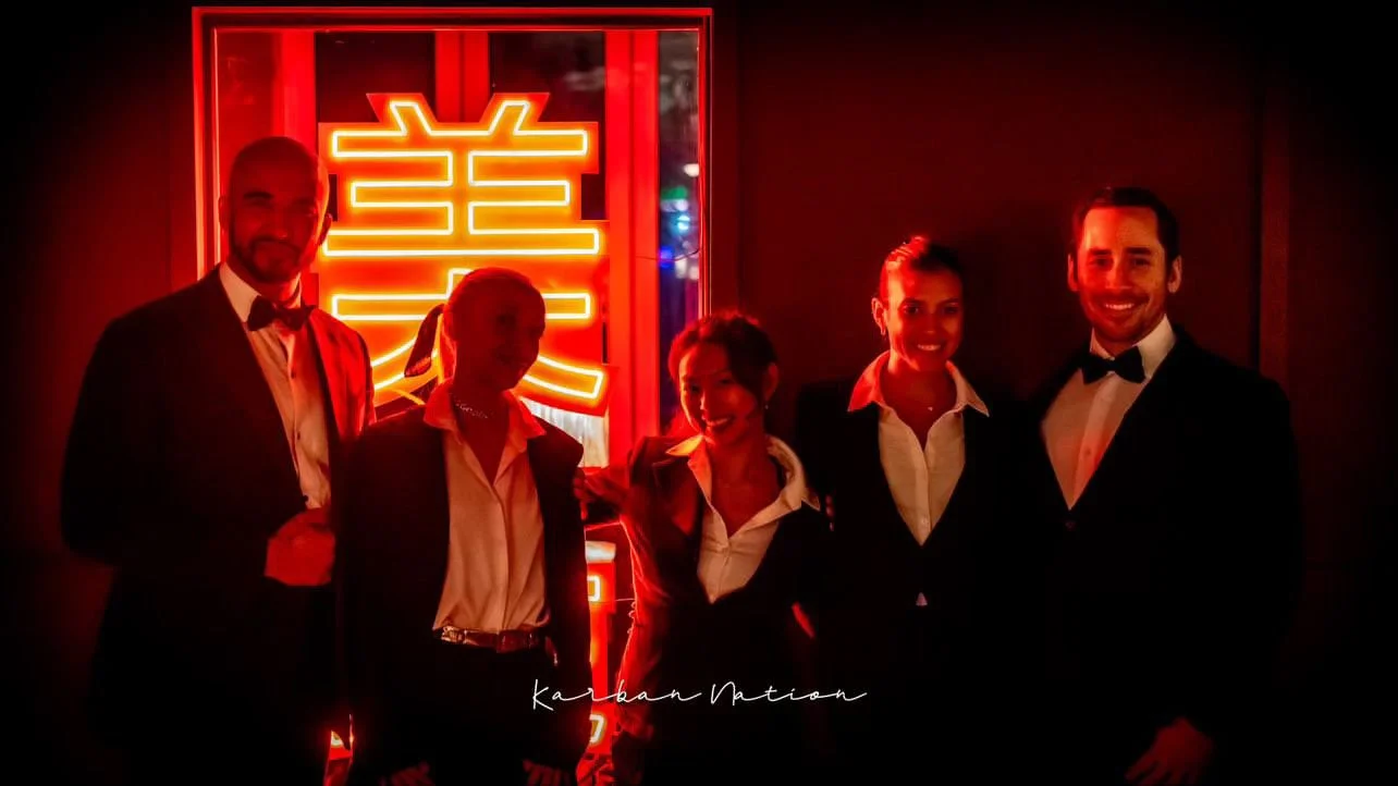 Five people dressed in formal tuxedos and suits posing in front of a neon red sign with Chinese characters in a dimly lit setting.