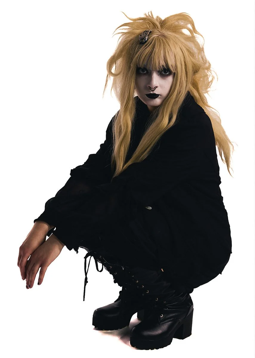 A person with long, messy blonde hair, wearing dark makeup, a septum piercing, and black lipstick, dressed in black clothing and black platform boots, squatting against a plain white background.