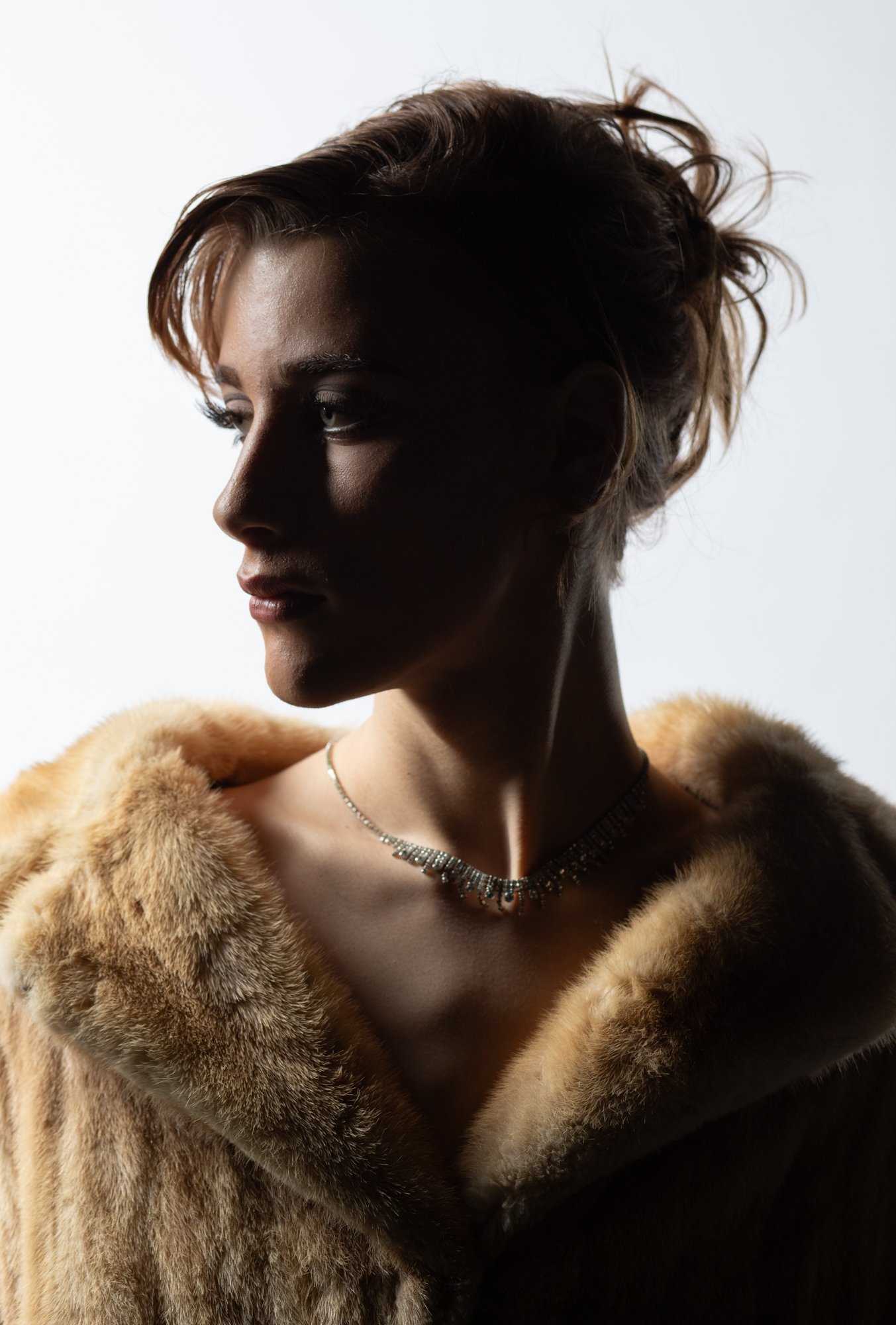 Side profile of a woman with short hair wearing a fur coat and a sparkly necklace, with a light background.
