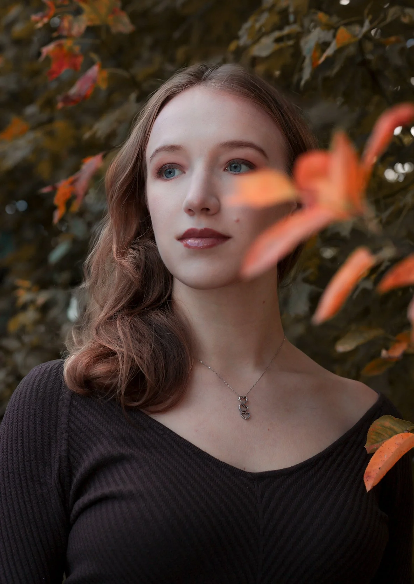 A young woman with long, wavy brown hair and blue eyes, wearing a black top and a necklace with four interlinked hearts, standing outdoors among orange and green autumn leaves.