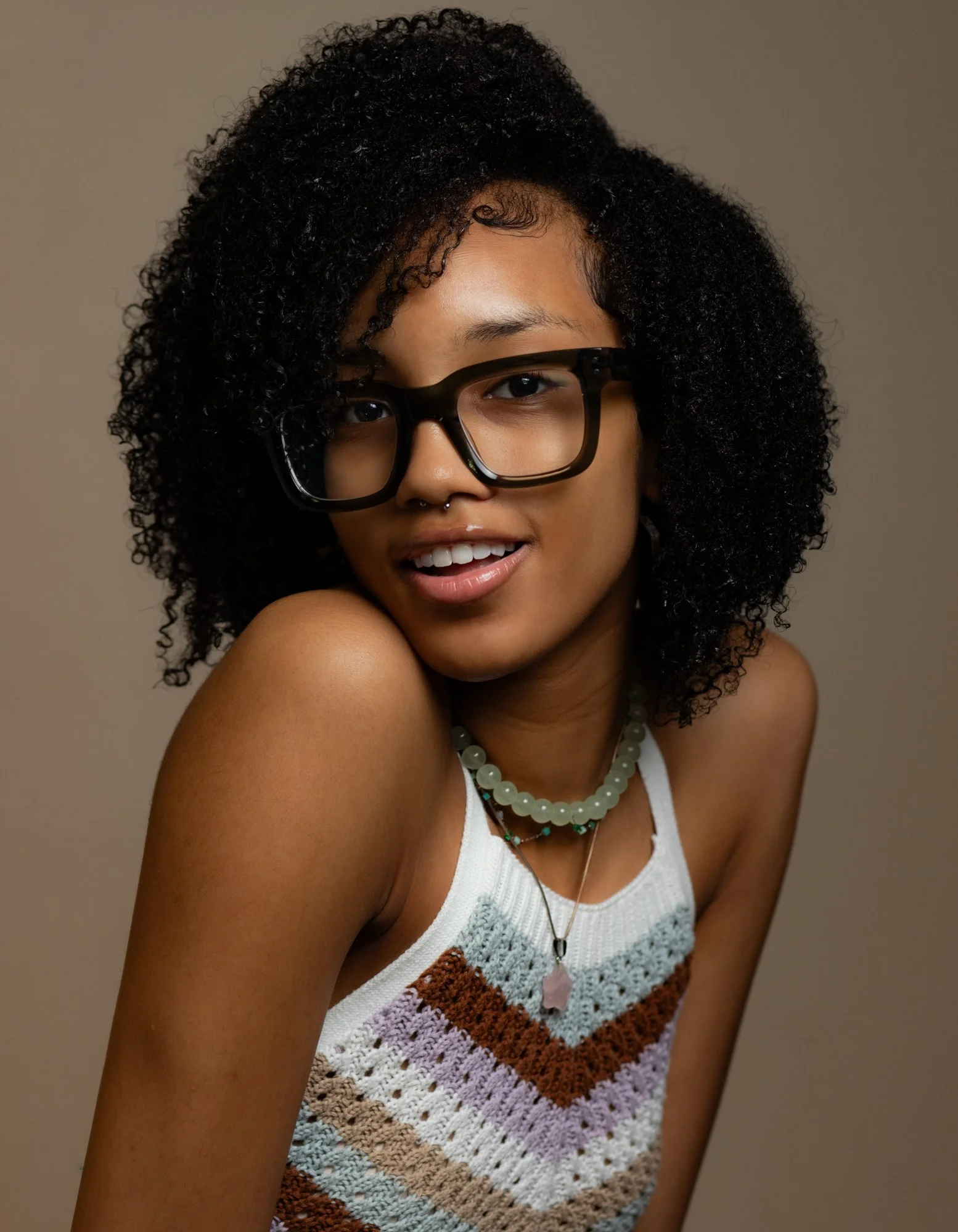 A young woman with curly black hair wearing large black glasses, a nose septum piercing, and a colorful crocheted tank top. She has layered necklaces and is smiling at the camera.