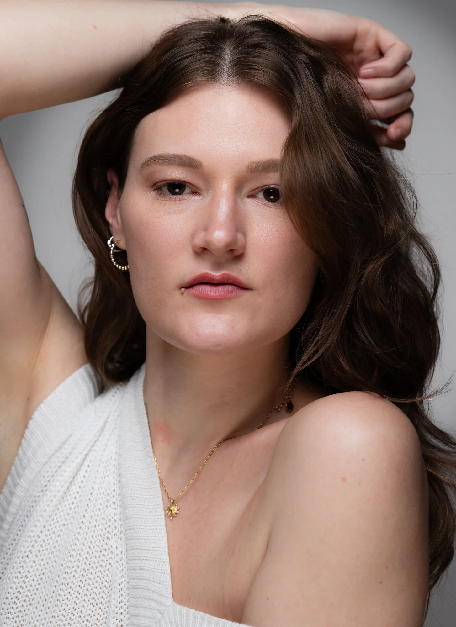 A close-up portrait of a woman with brown, wavy hair, one arm raised behind her head, wearing hoop earrings and a gold necklace with a flower pendant, dressed in a white knit top, against a neutral background.