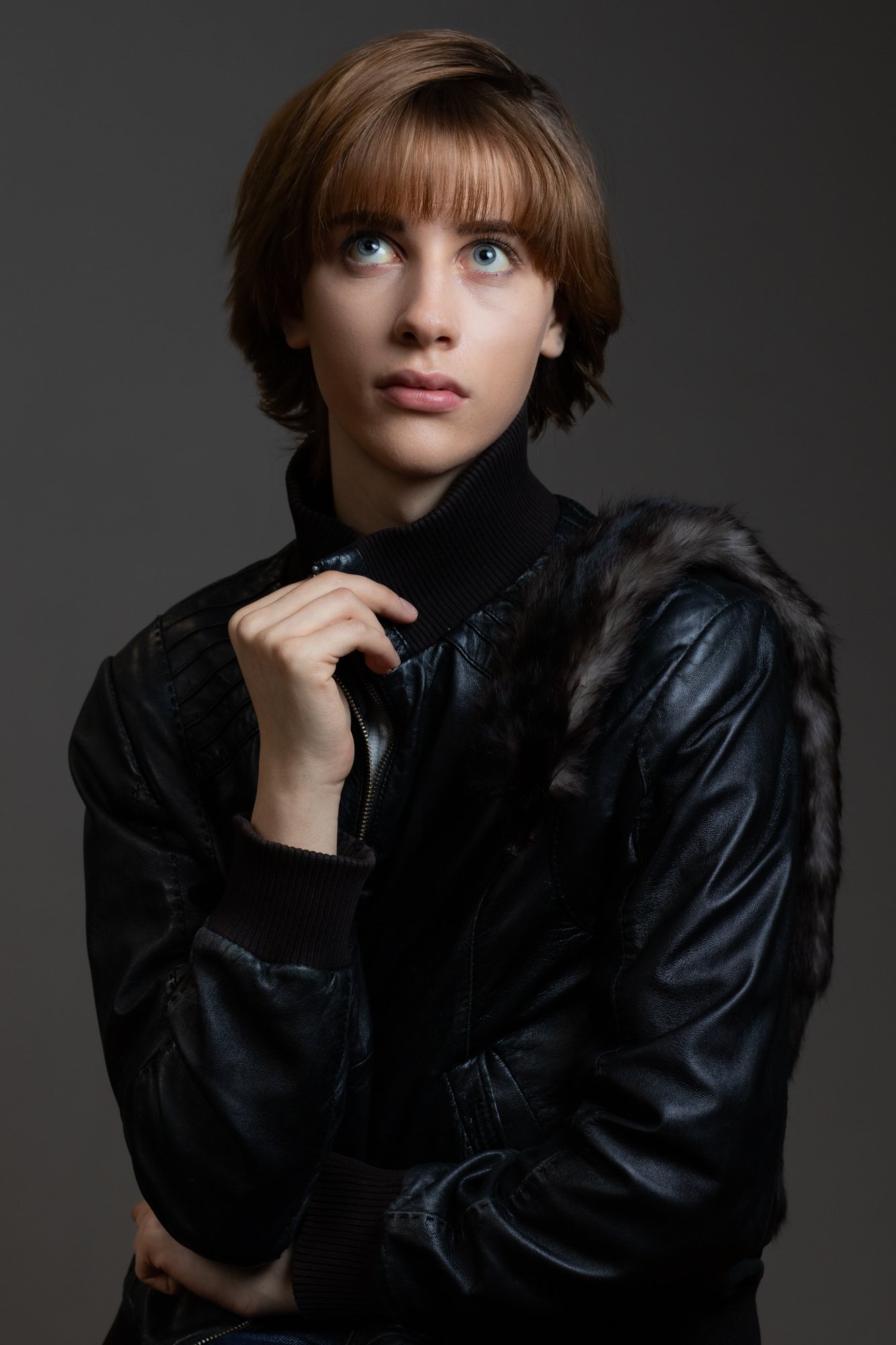 Portrait of a young woman with short brown hair and blue eyes, wearing a black leather jacket with faux fur collar, against a dark background.