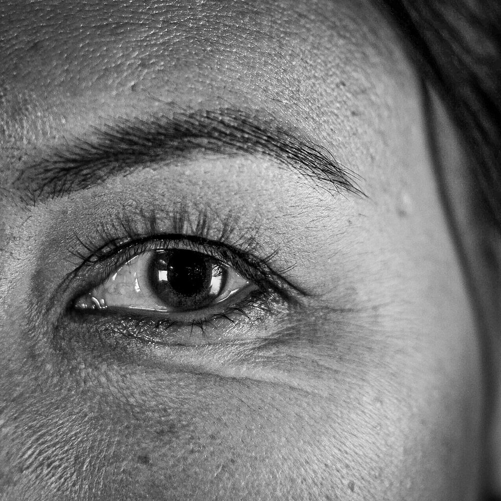 Close-up black-and-white photo of a human eye, showing detailed skin texture around the eye, eyebrow, and eyelashes.