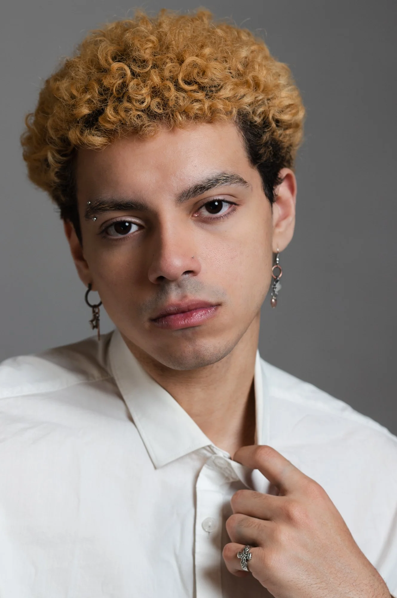 Close-up portrait of a young person with curly blond hair, wearing earrings, and a white shirt. They are holding the collar of their shirt with one hand.
