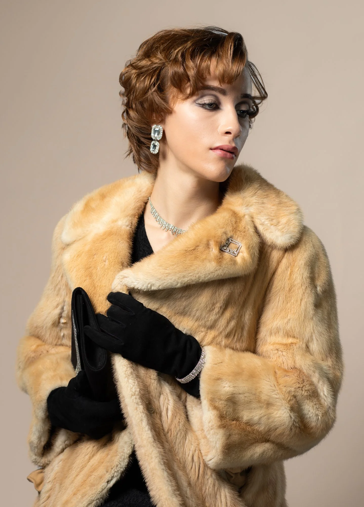 Woman with short, wavy auburn hair wearing a beige fur coat, black gloves, and diamond jewelry, posing against a neutral background.