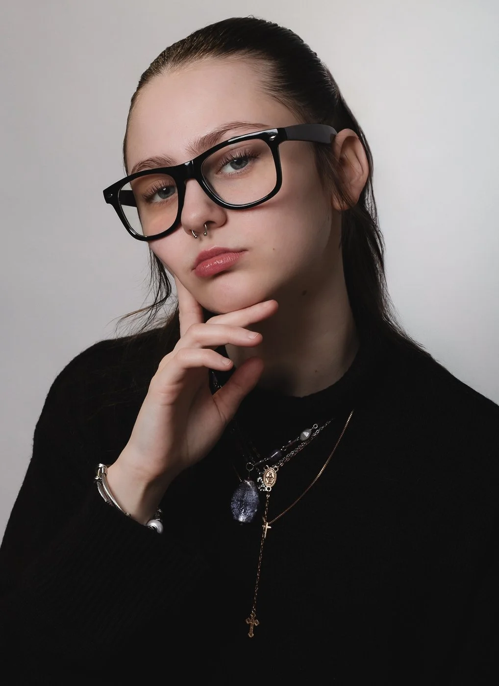 A young woman with glasses, piercings, and layered necklaces posing with her hand on her chin.