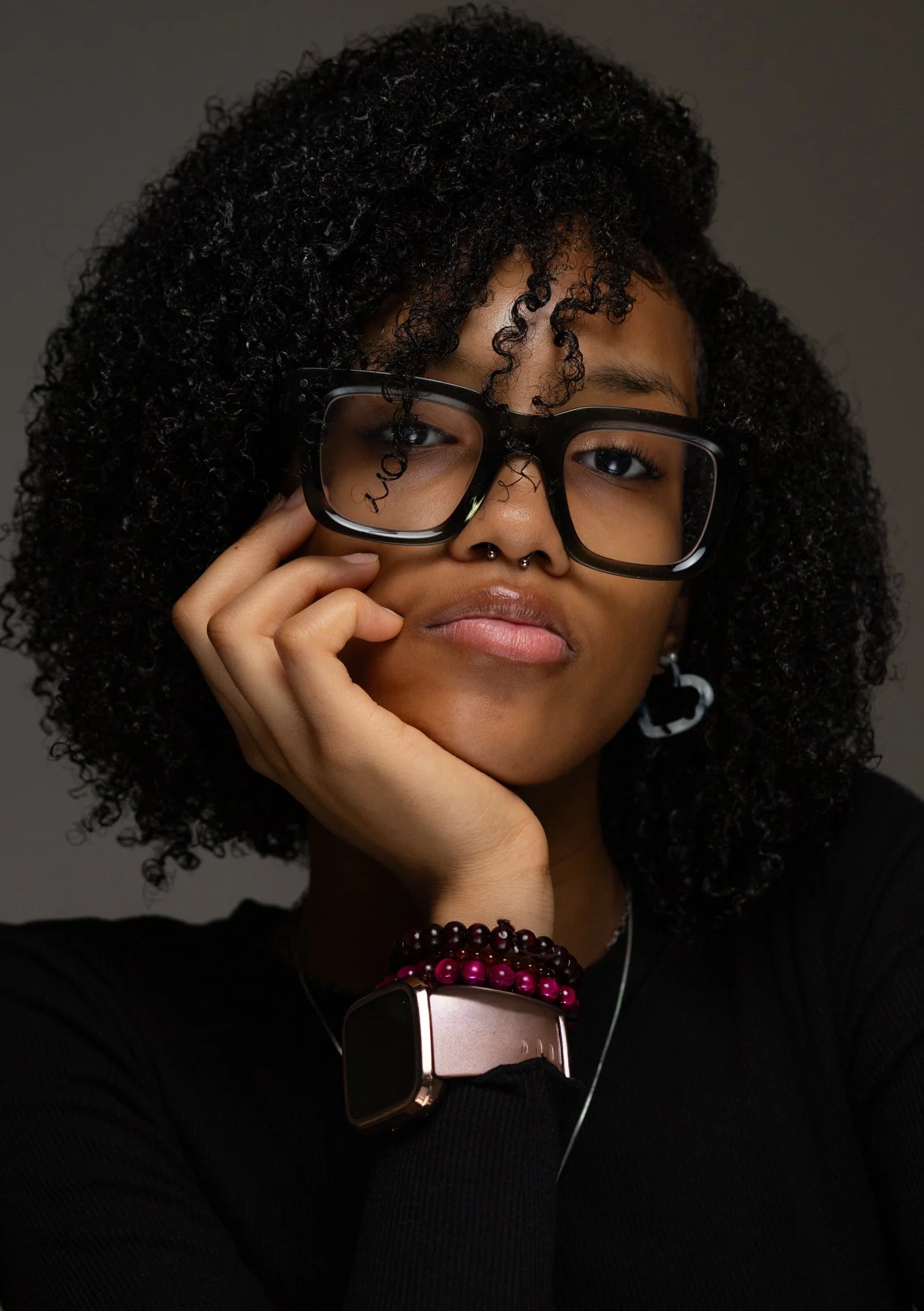 A woman with curly hair wearing large black glasses, a smartwatch, and beaded bracelets, resting her face on her hand.