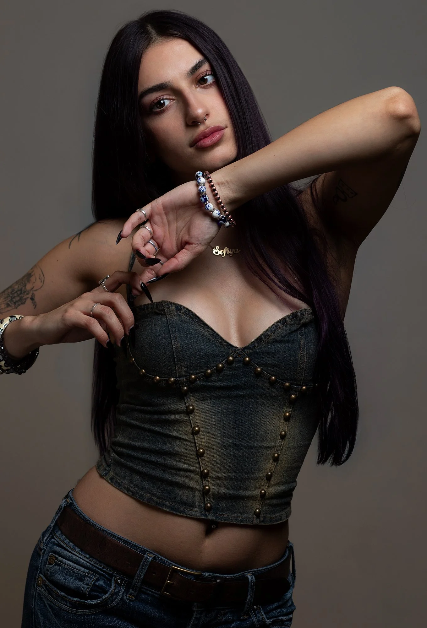 Young woman with dark hair and tattoos posing against a neutral background, wearing a strapless denim top, jeans, and various jewelry including bracelets, rings, and a necklace.