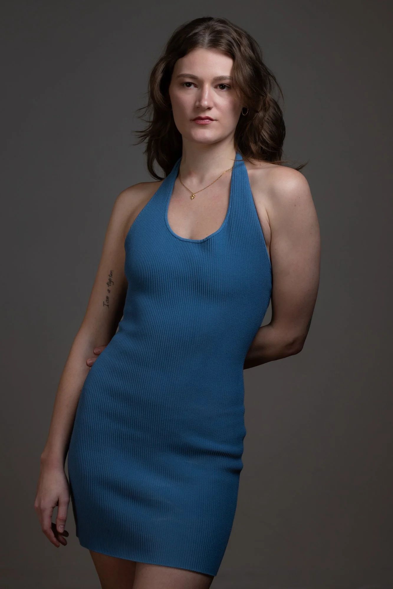 A woman with brown wavy hair wearing a blue sleeveless dress standing against a gray background.