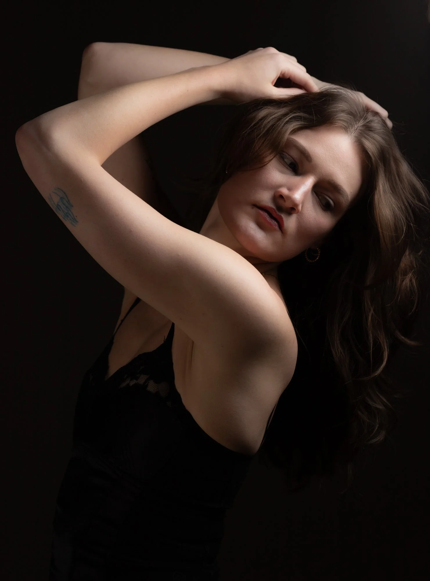 A woman with wavy brown hair posing against a dark background, wearing a black lace top, with her arms raised and hands touching her head.