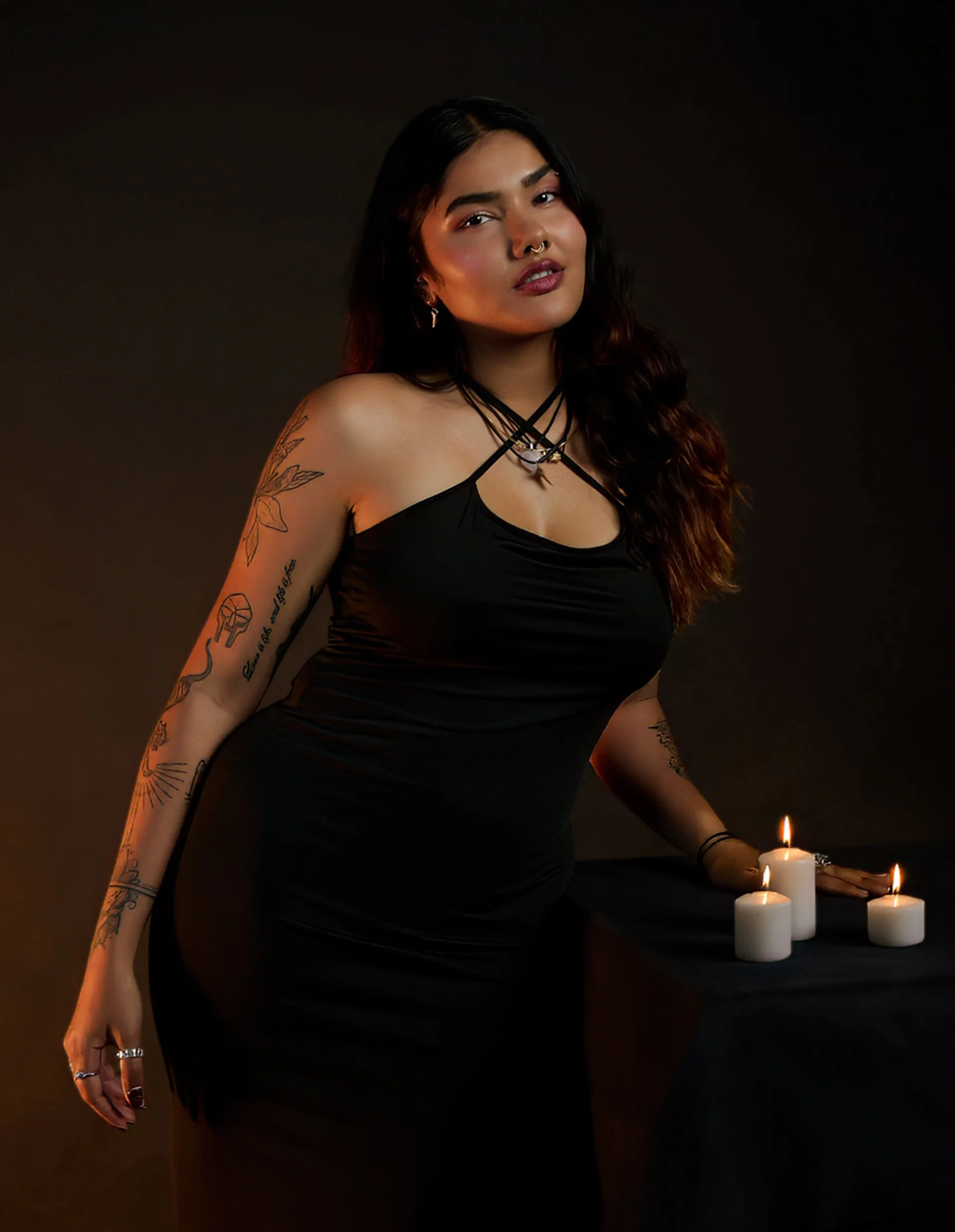 Woman with dark hair and tattoos wearing a black dress, standing next to a table with white candles, in a dark setting.