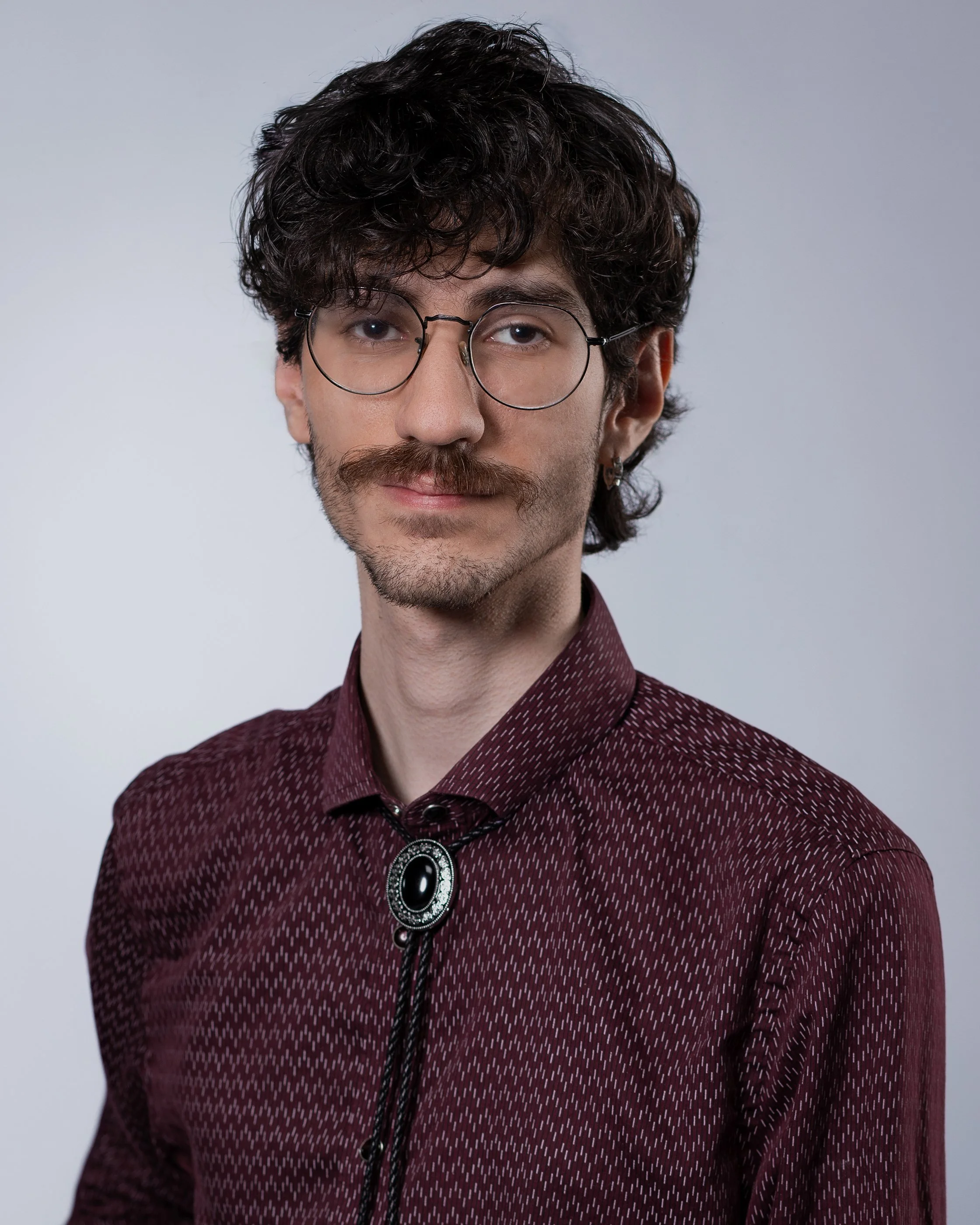 A young man with curly dark hair, glasses, and a mustache, wearing a maroon shirt with a bolo tie, posing against a plain background.