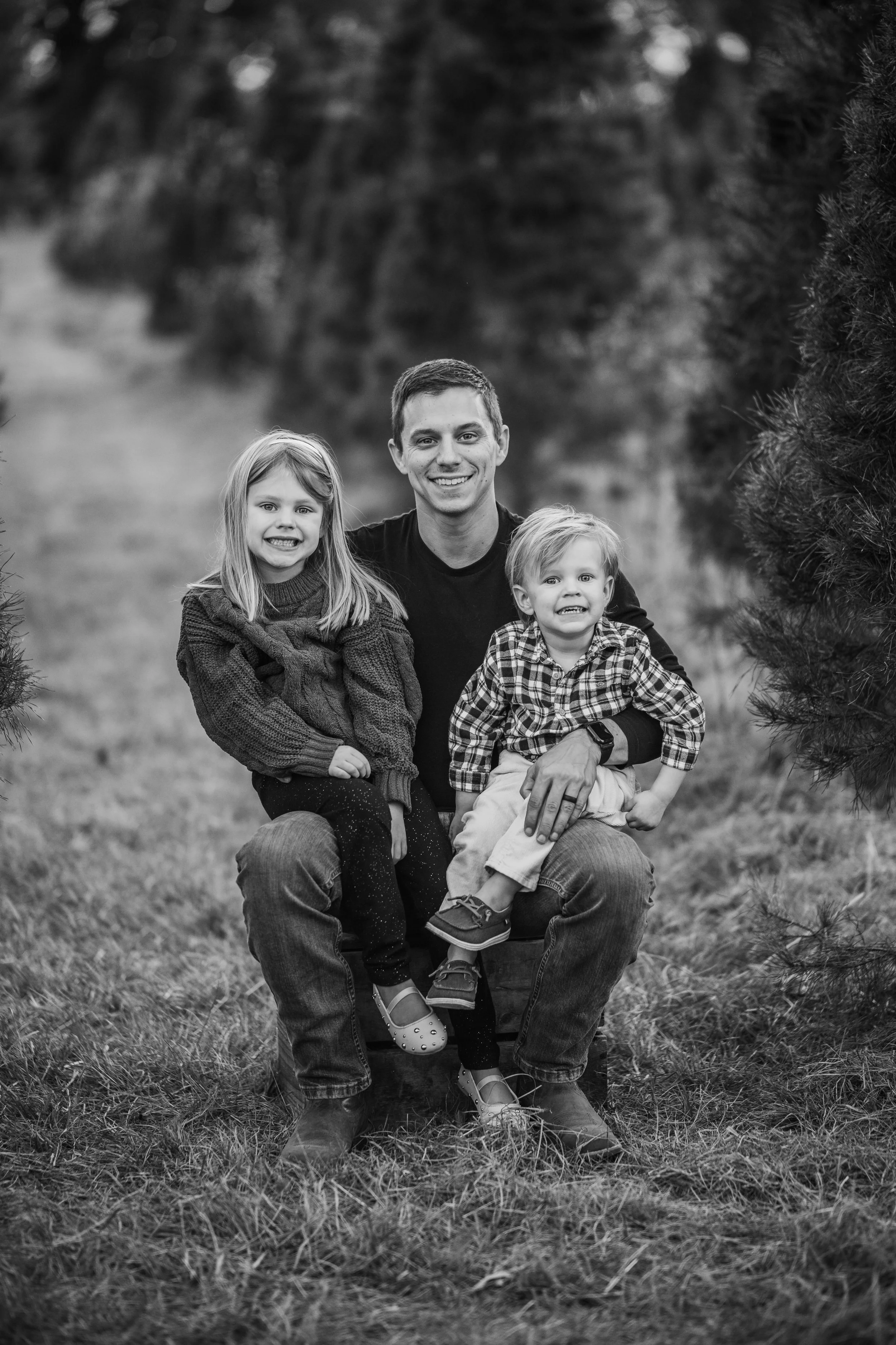 A black and white photo of a man with two children on his lap outdoors, all smiling, surrounded by trees.