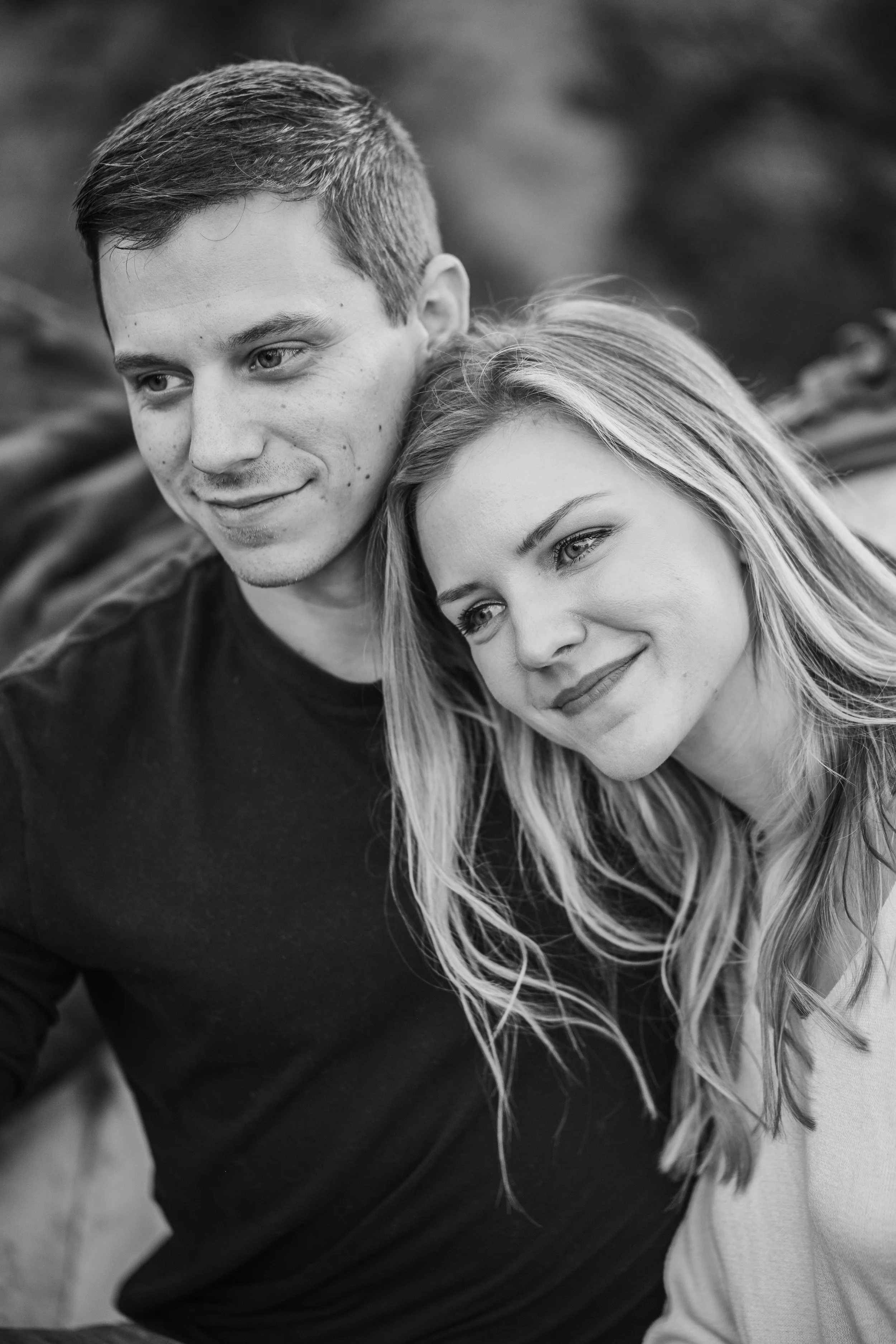 A close-up black and white photo of a young man and woman smiling and sitting close together outdoors.