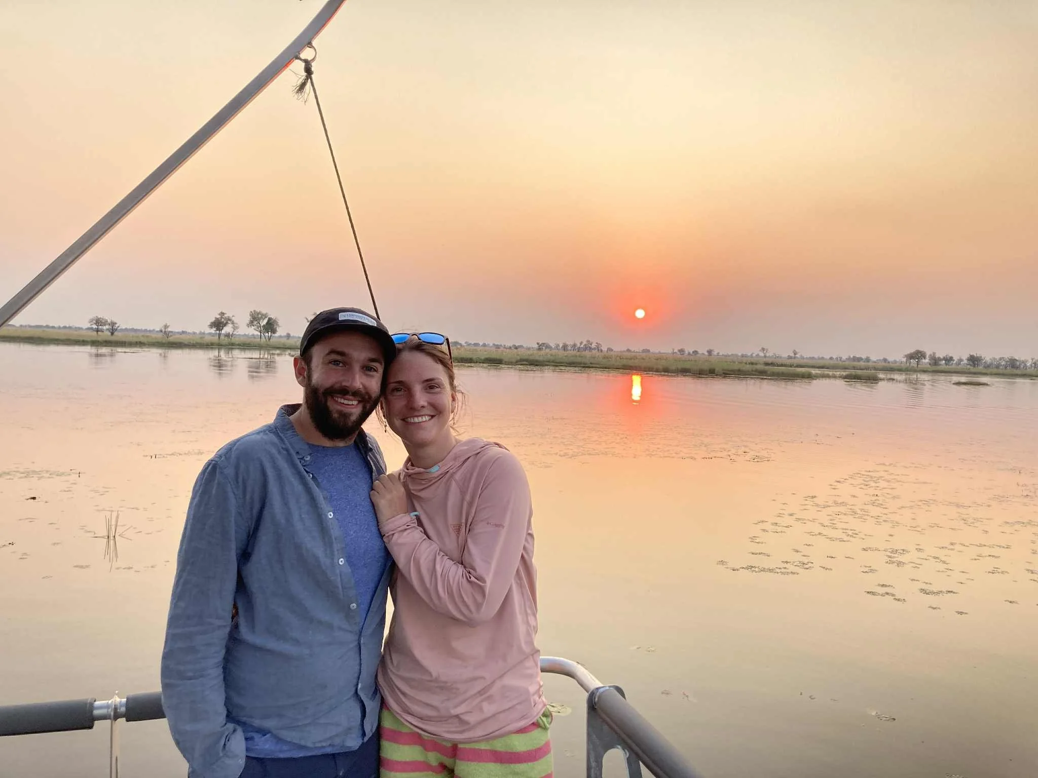 A happy couple standing on a boat at sunset, smiling, with a calm river and distant trees in the background.