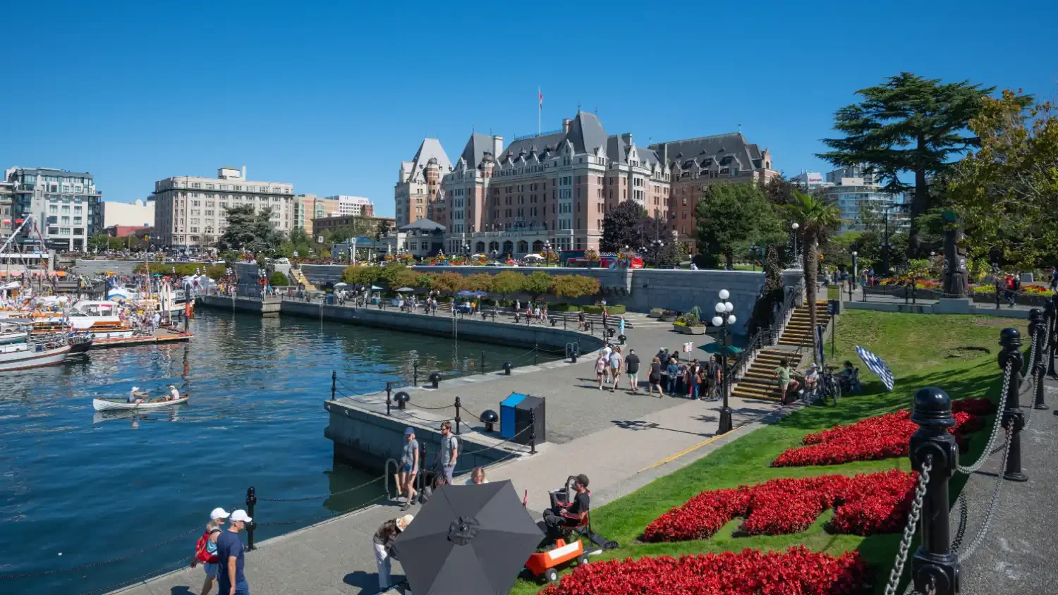 View of a waterfront promenade with boats, people walking, trees, and a large historical hotel building in the background on a sunny day.
