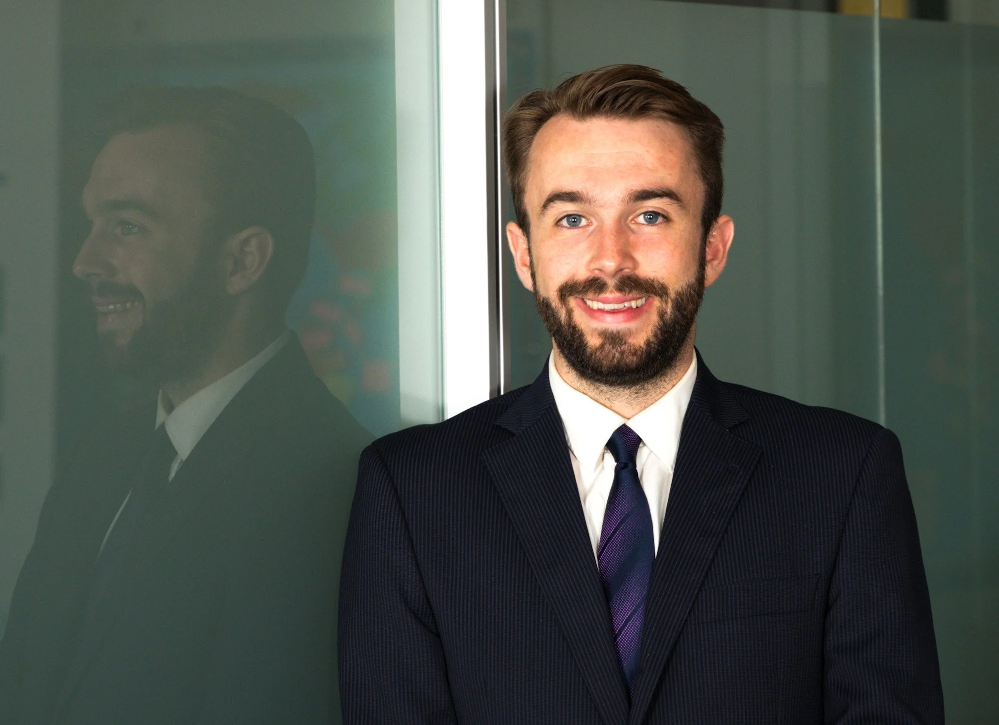 A smiling man with a beard and light brown hair in a business suit with a striped tie, standing next to a glass wall that reflects his face.